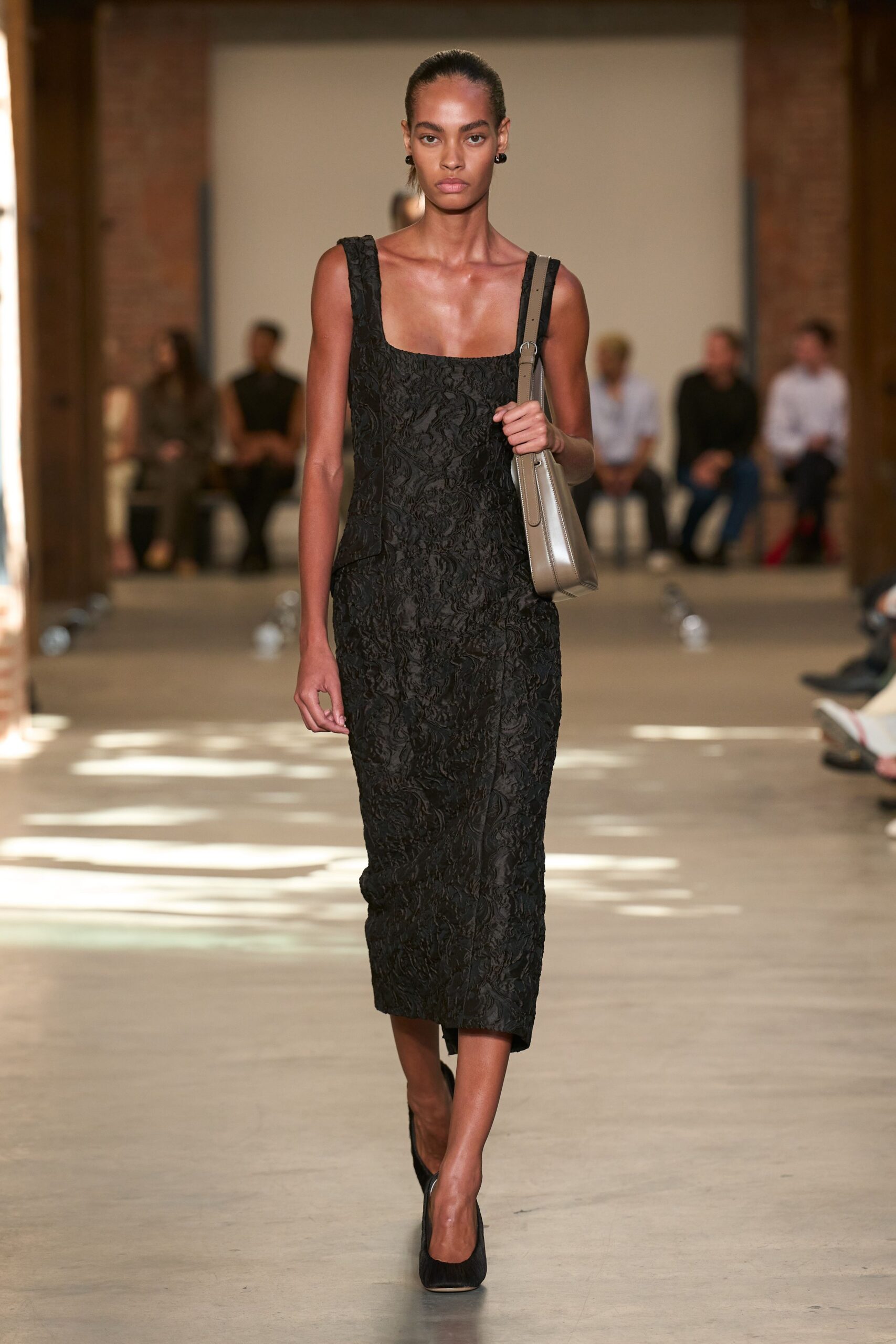 Black square-neck midi dress in crushed floral jacquard, one hip slightly kicked out. Taupe leather shoulder bag with a long tabbed seam. Black pumps in a matte woven finish. Walking into a hotel bar too early for dinner, sunlight still on the floor.