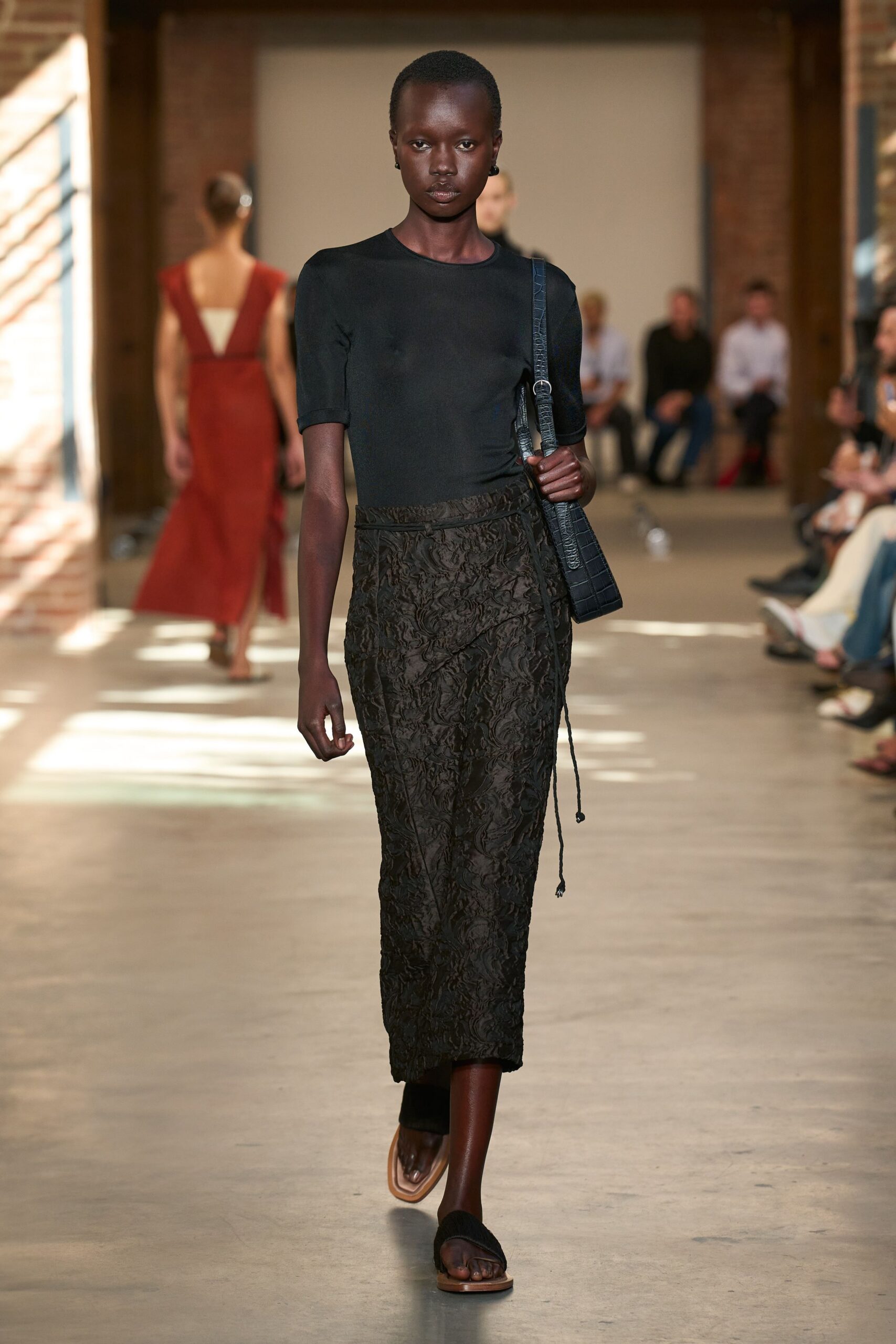 Black stretch tee with elbow sleeves and a close crew neck. Black crinkled jacquard wrap skirt, cords hanging loose at the hip. Embossed black shoulder bag with a narrow crocodile strap. Flat black thong sandals with pale leather soles. For crossing a sunstruck courtyard after lunch ran two hours long.