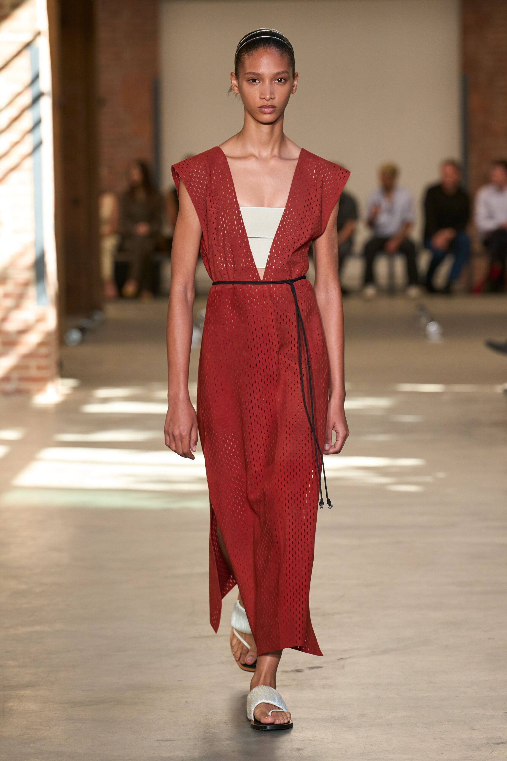 Brick red perforated dress with broad cap shoulders and a plunging V. Chalk bandeau inset, one narrow black seam across the front. Black cord belt tied low, twin ends dangling past the hip. White hairy slide sandals on flat black soles. Crossing a concrete courtyard just after the heat starts to lift.