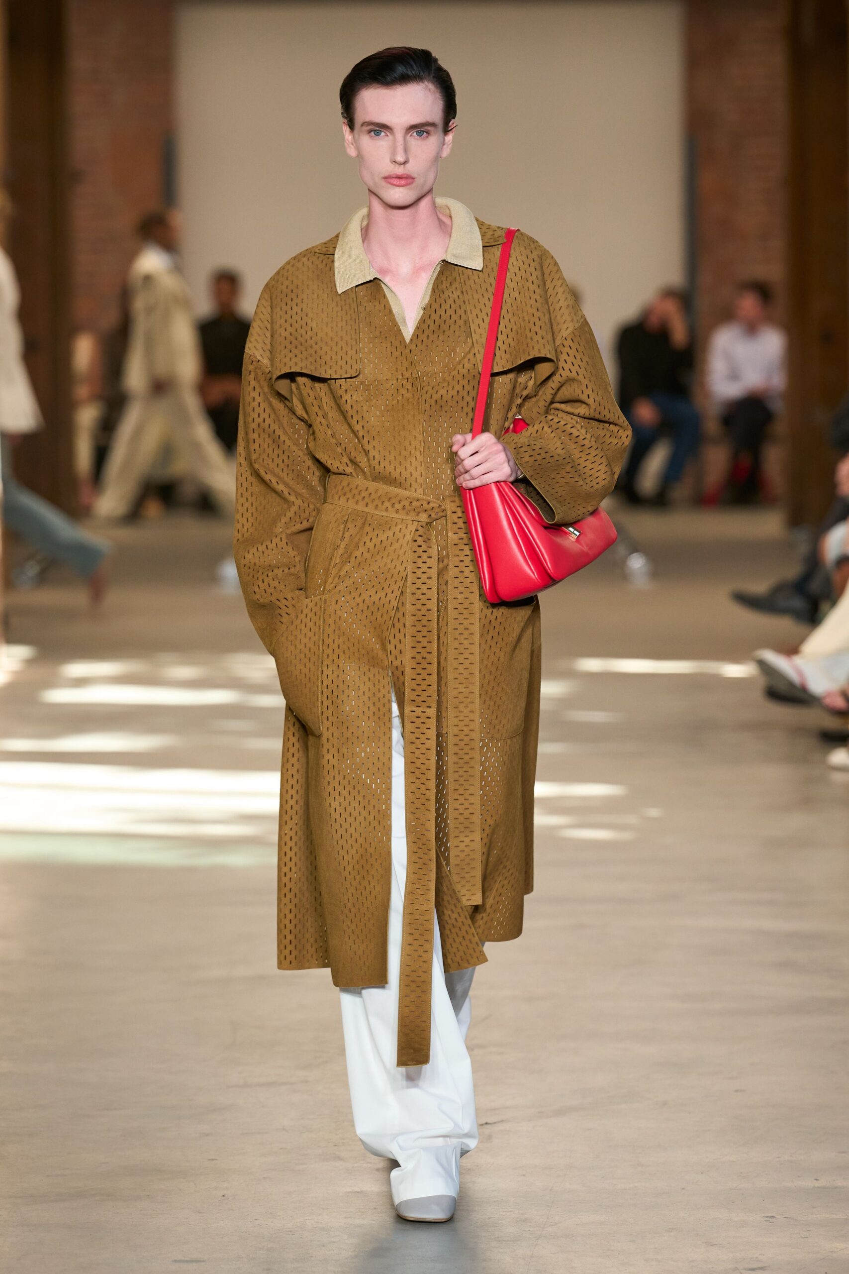Ochre laser-cut leather coat with a pale felt collar and long self-belt. White wide-leg trousers breaking softly over the shoe. Tomato red leather shoulder bag, padded into folded ridges. Square-toe white boots with a dove-grey cap. Crossing a museum atrium while the rain still dries on the stone.