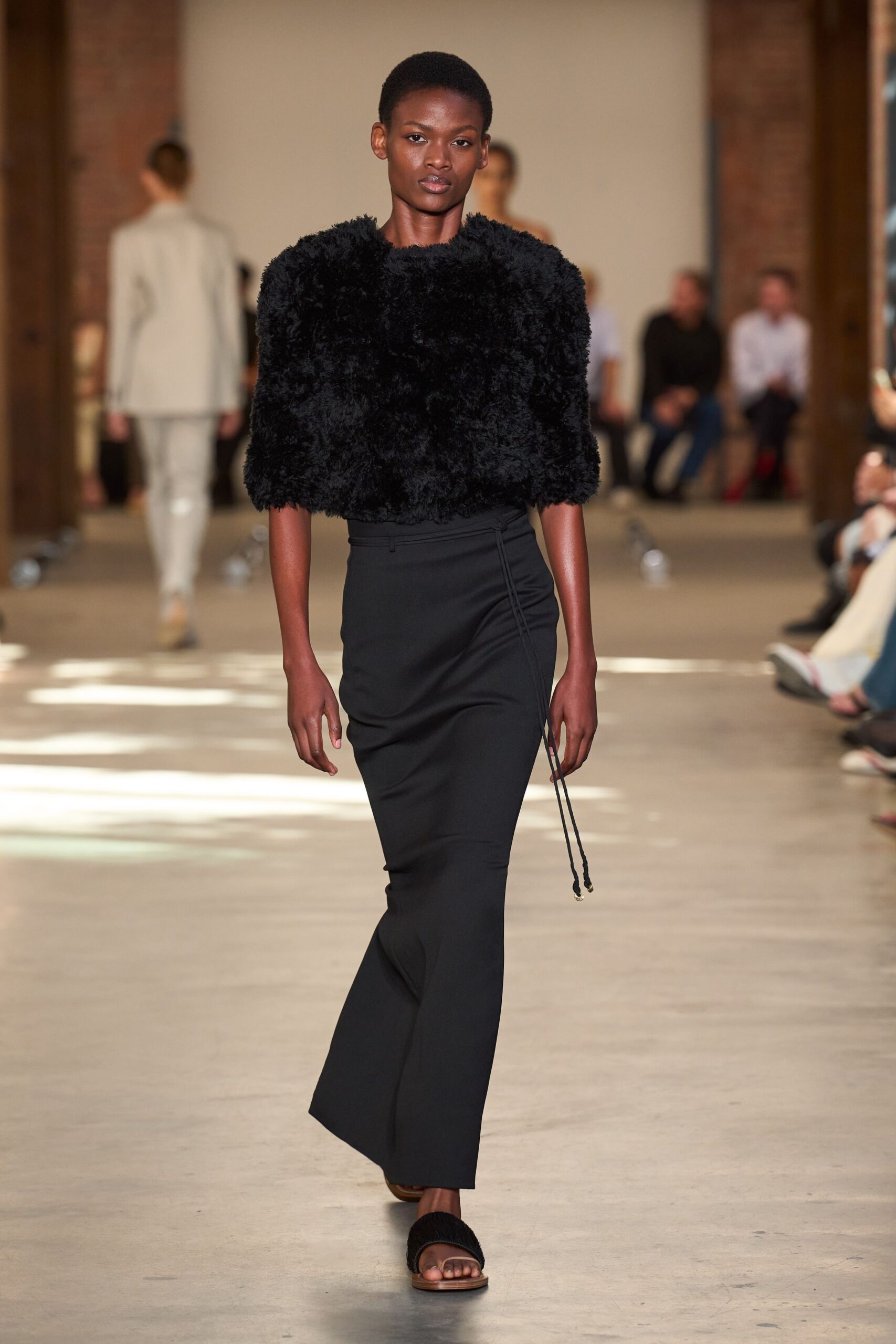 Black shearling top cropped at the waist, sleeves ending below the elbow. Column skirt in matte black crepe, wrapped with a cord tie tipped in metal. Flat sandals with shaggy black straps. For crossing a silent concrete lobby before the building fully wakes.