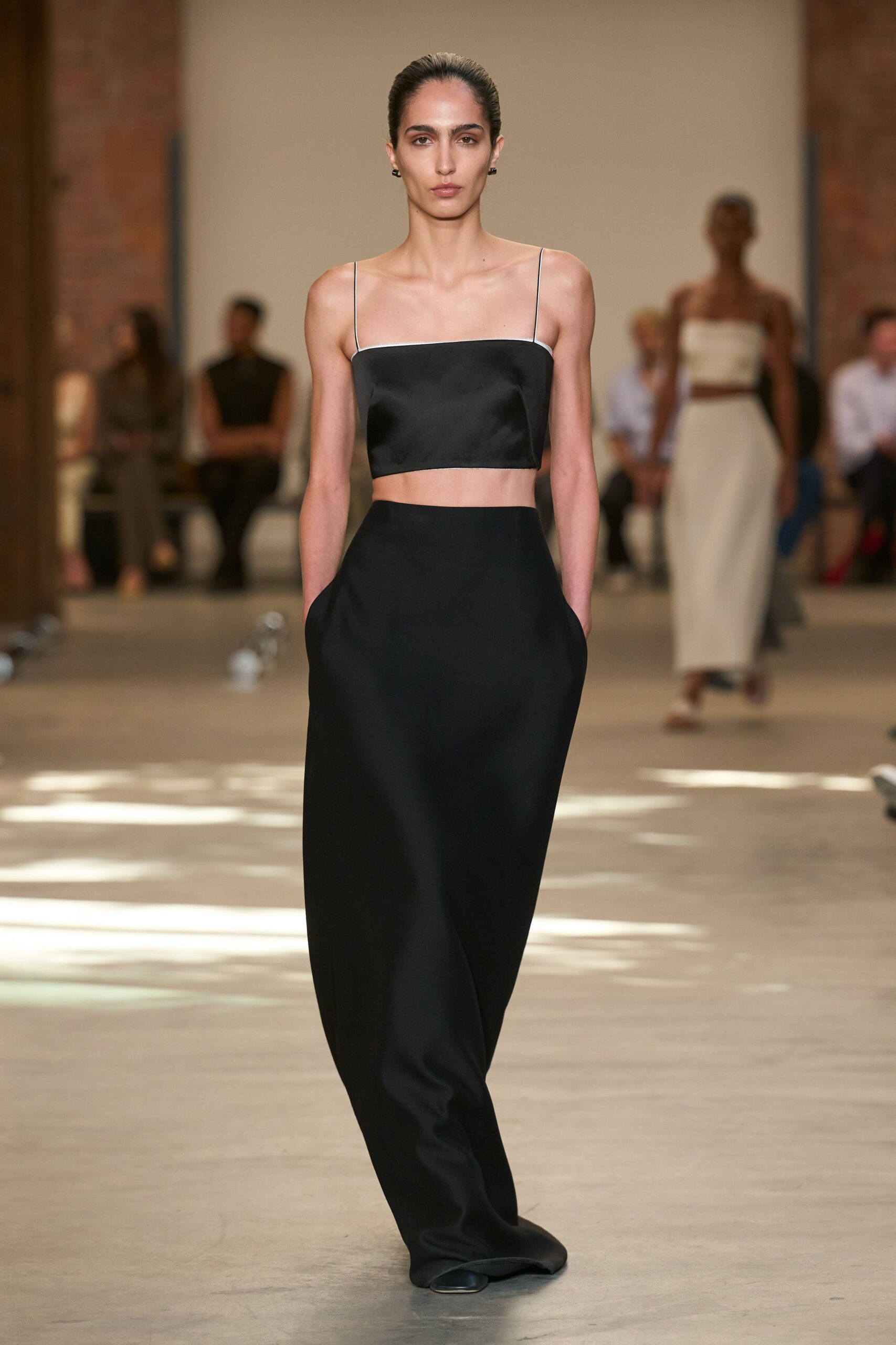 Black satin crop top with white piped edge and threadlike straps. Black column maxi skirt, pocketed at the hips, hem dragging over square toes. Small black studs. Morning after a hotel wedding, crossing the lobby barefoot in your head.