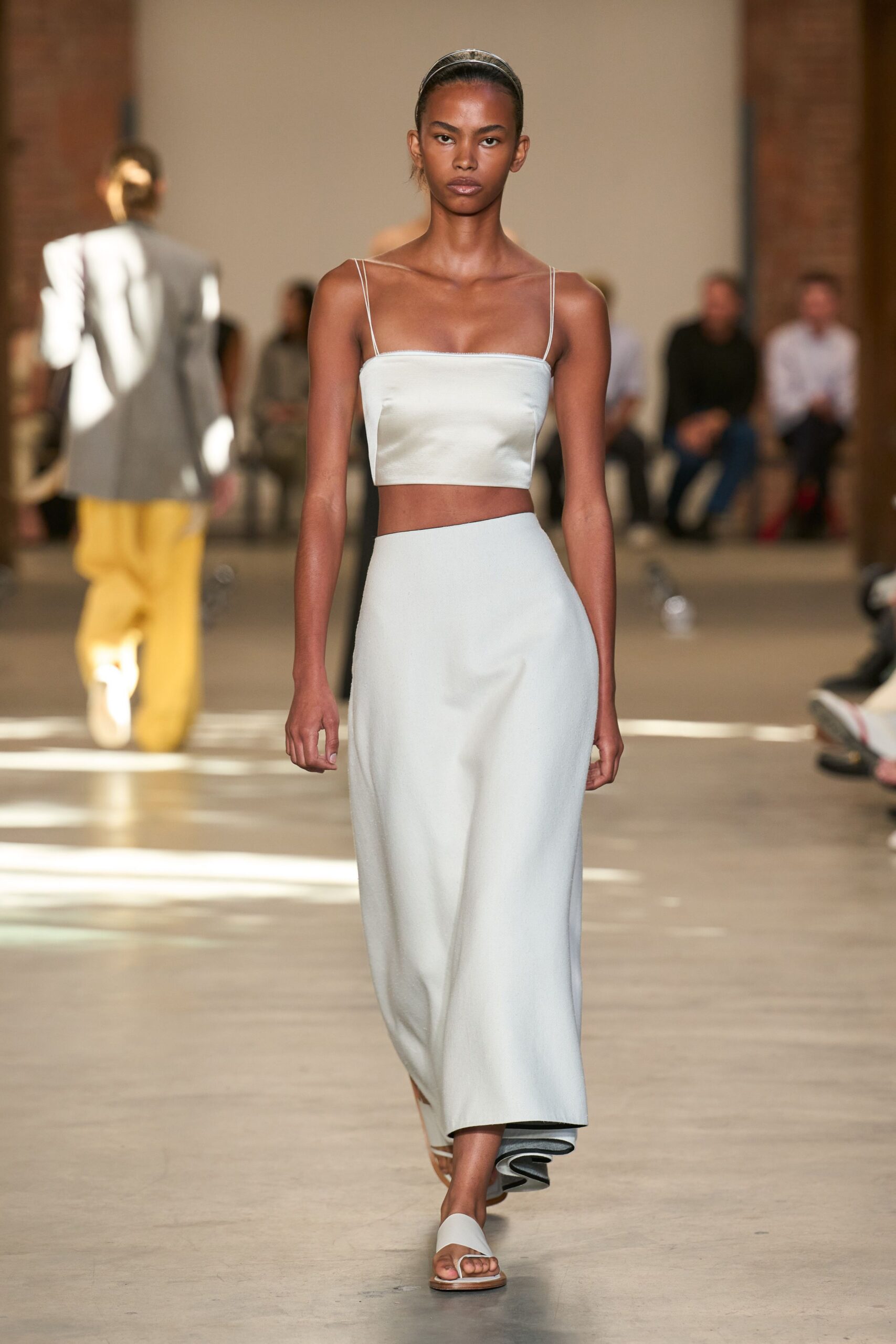 Narrow metallic headband holding the hair flat off the face. Pale blue satin crop top with doubled spaghetti straps. Bias-cut midi skirt in the same satin, hem curling darker underneath. White slide sandals with a flat tan sole. Walking back from the pool bar before the terrace tables fill for dinner.