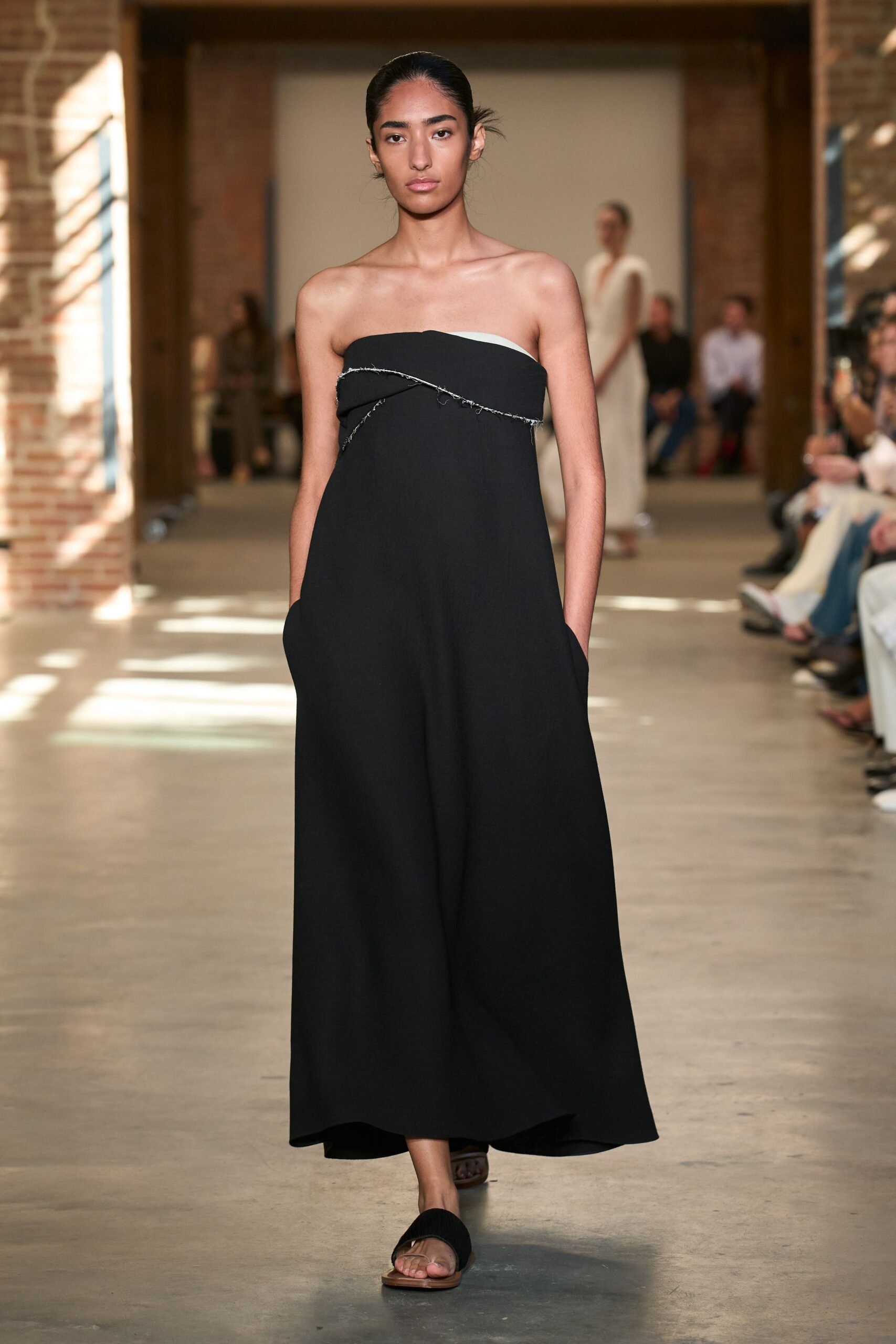 Black strapless column dress with a slanted raw-edged band exposing white underneath. Black wide-strap flat sandals on thin tan soles. Crossing a sunstruck cloister after the speeches are finally over.