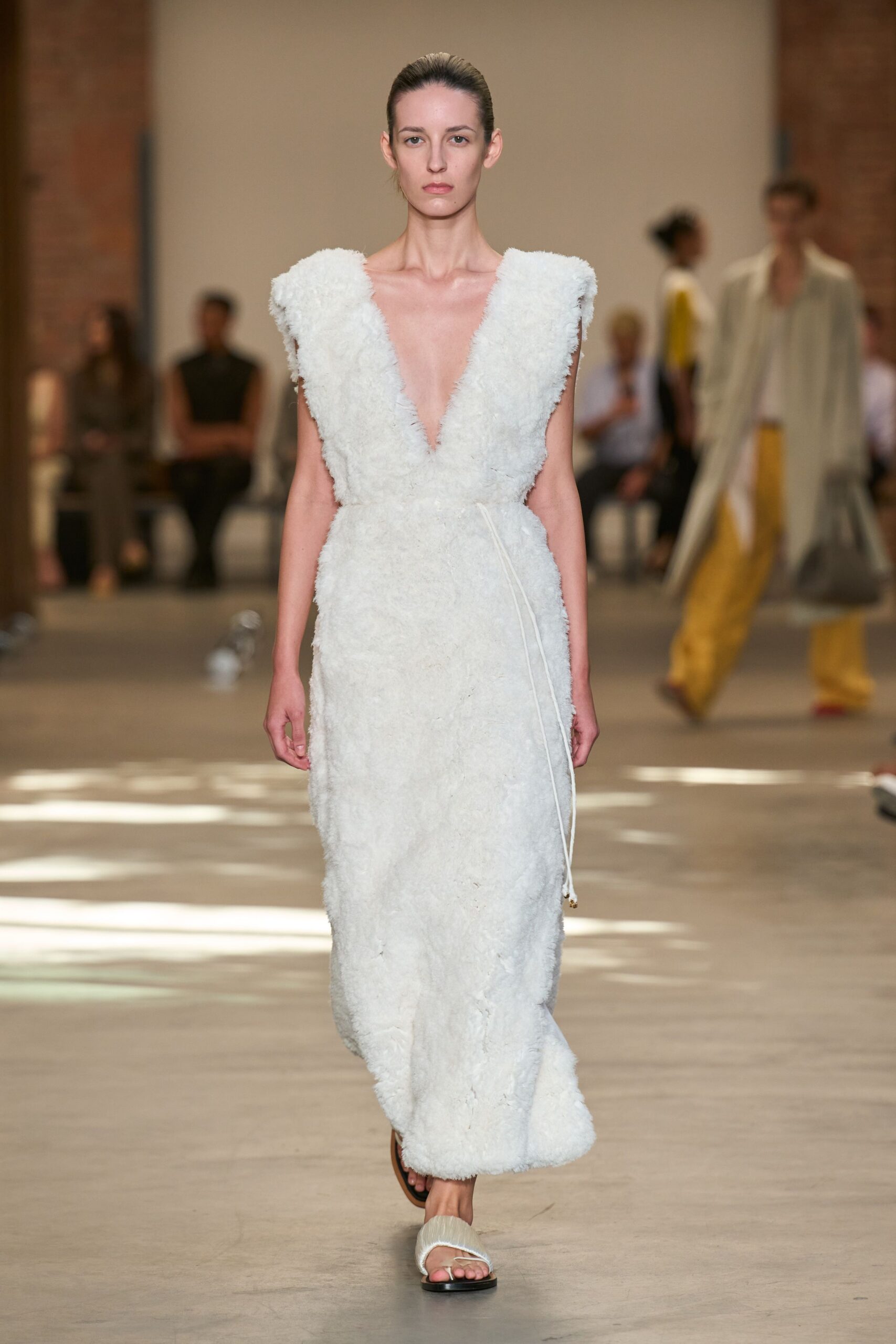 Ivory shag-pile column dress with a plunging V and cord tie dangling loose. Padded white thong sandals on flat black soles. Crossing a quiet spa lobby before anyone else is awake.