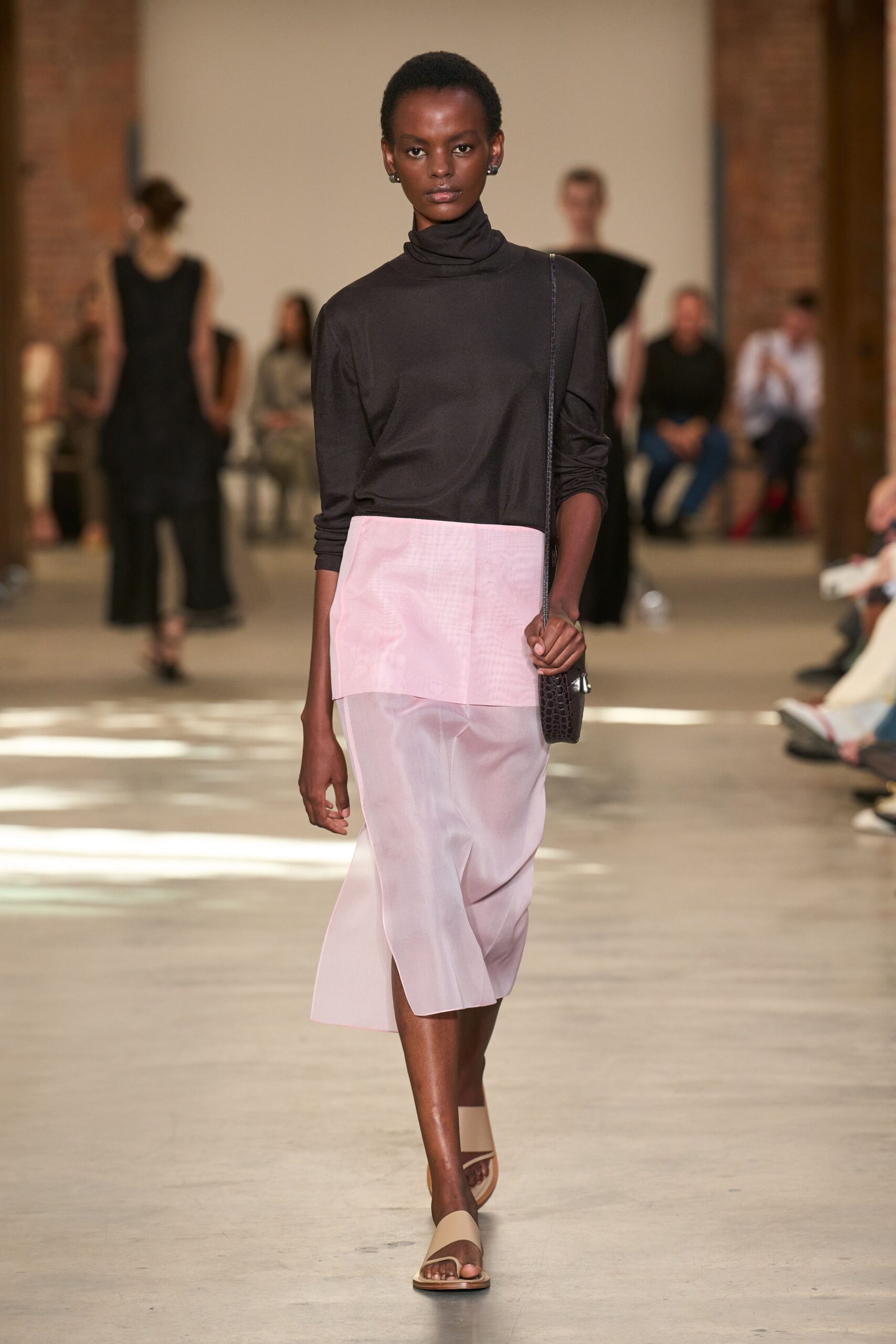Black jersey turtleneck with a loose rolled neck and pushed sleeves. Pale shell-pink organza skirt, sheer below an opaque hip panel. Slim crocodile-embossed pouch hanging on a narrow strap. Nude leather toe-ring sandals with a square-cut front strap. Walking through a museum atrium before the doors open to the heat.