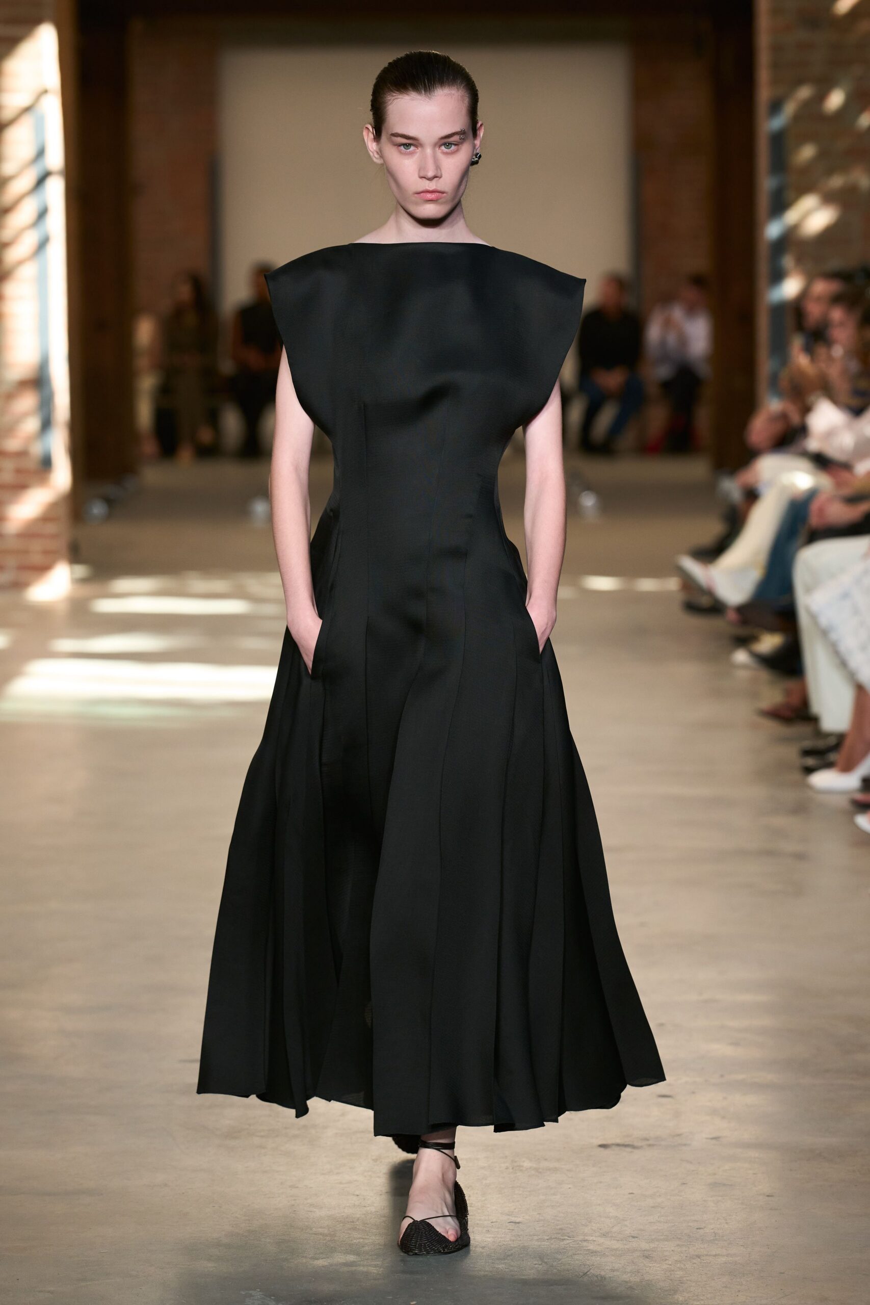 Black structured gown with a boatneck yoke and deep side pockets. Black woven thong sandals with slim ankle ties. Dark sculptural ear cuff hugging one lobe. Crossing a museum atrium before the sound check starts.