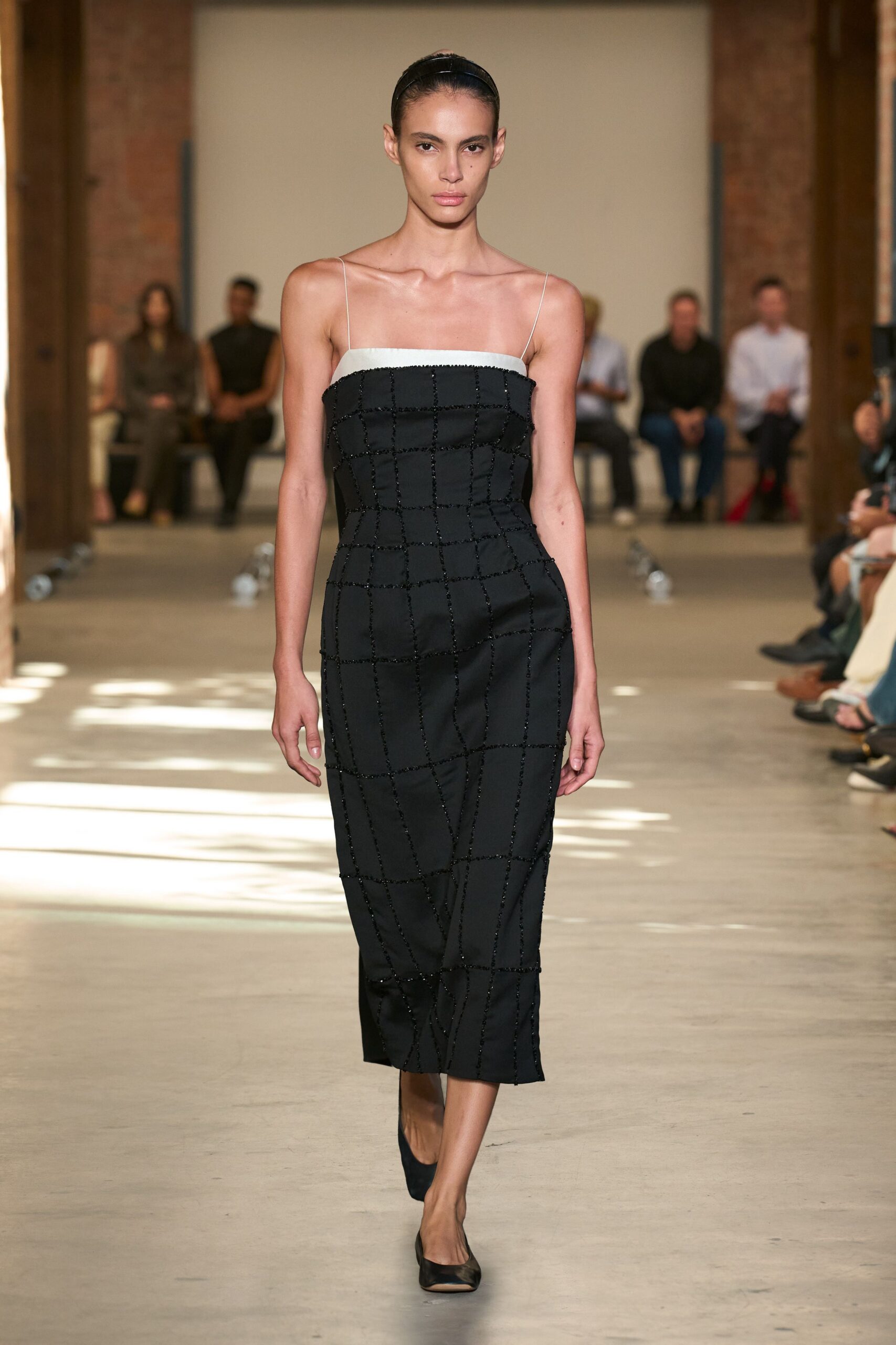 Narrow black satin headband, hair pulled back flat beneath it. Ivory-trimmed column dress on filament straps, black beaded grid buckling off straight lines. Black pumps with a low curved vamp. Crossing a museum atrium just after the school groups cleared out.