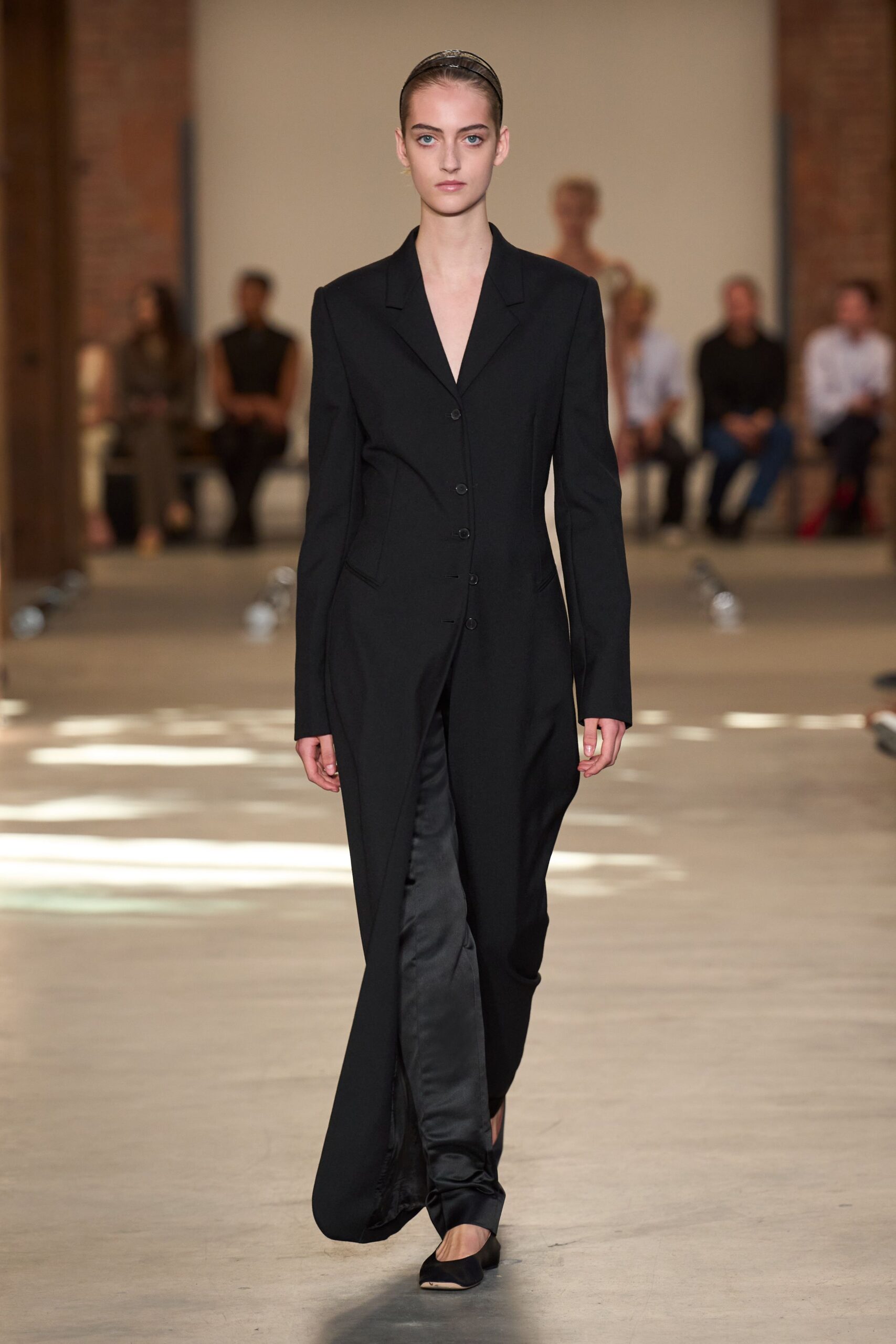 Narrow black headband with a faint patent gleam. Long black tailoring coat, five small buttons and a dipped front opening. Black satin trousers pooling softly over the instep. Black pointed flats cut low across the foot. For crossing a museum atrium before anyone has unlocked the offices.