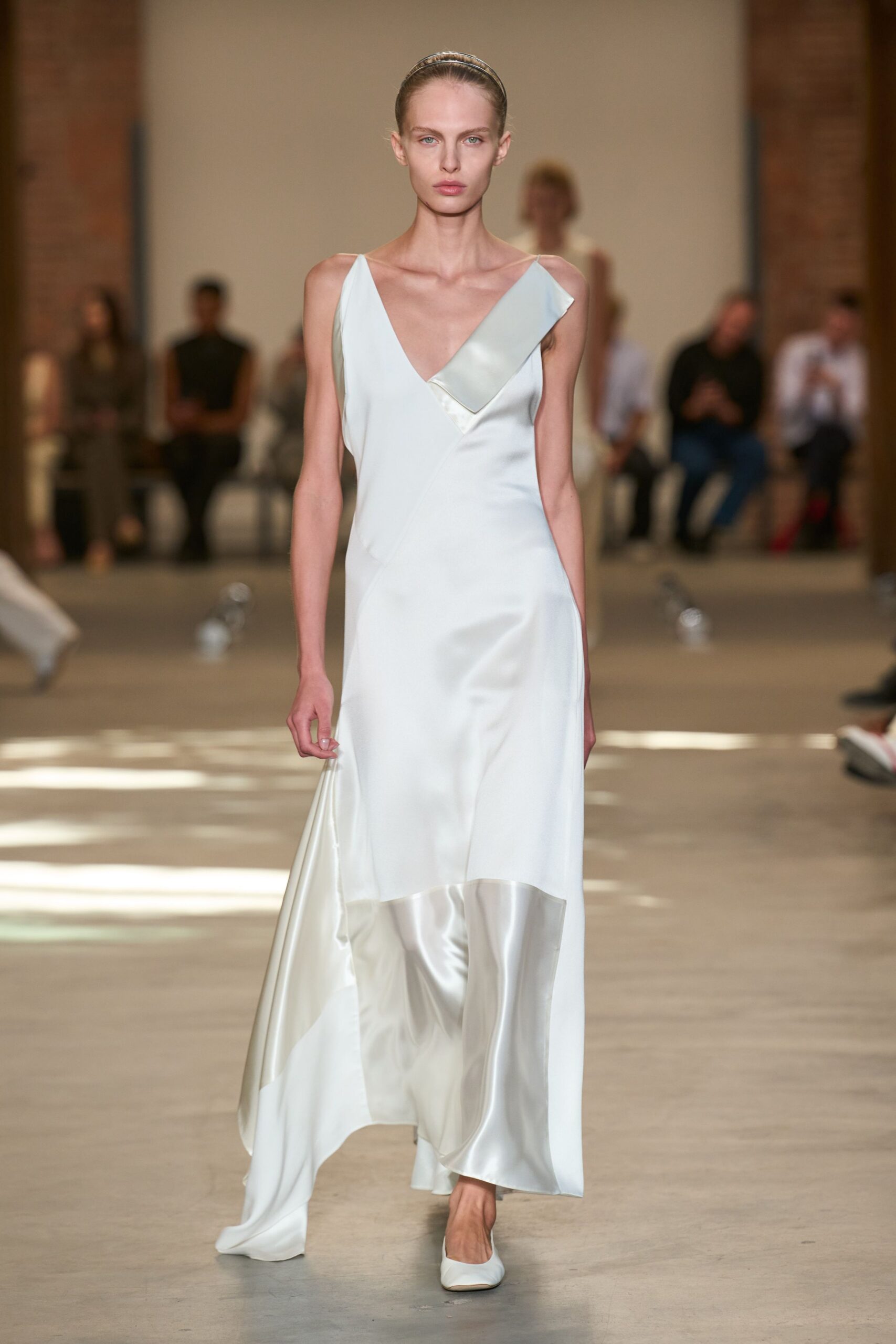 Slim metallic headband holding the hair flat off the face. Pale ice-blue satin slip dress with a folded bib crossing the deep V. White pumps with a softly squared toe. Crossing a hotel lobby at dawn, still carrying last night's calm.