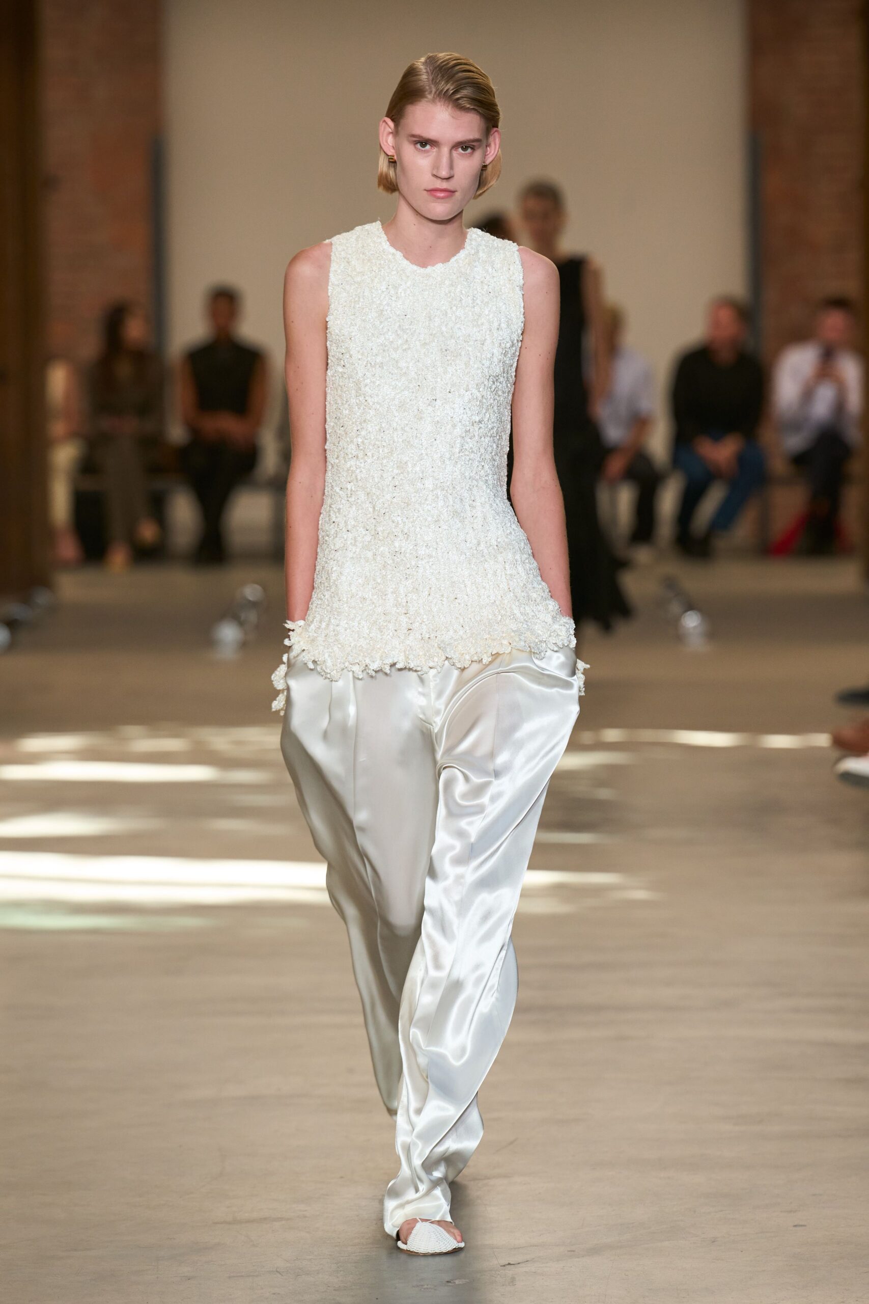 Ivory boucle shell, hand-frayed into little nubs along the hem. Pearly satin trousers cut loose, pooling in liquid folds over the foot. White mesh mules with a pointed peep toe. Morning after the wedding, carrying coffee across a tiled hotel corridor.
