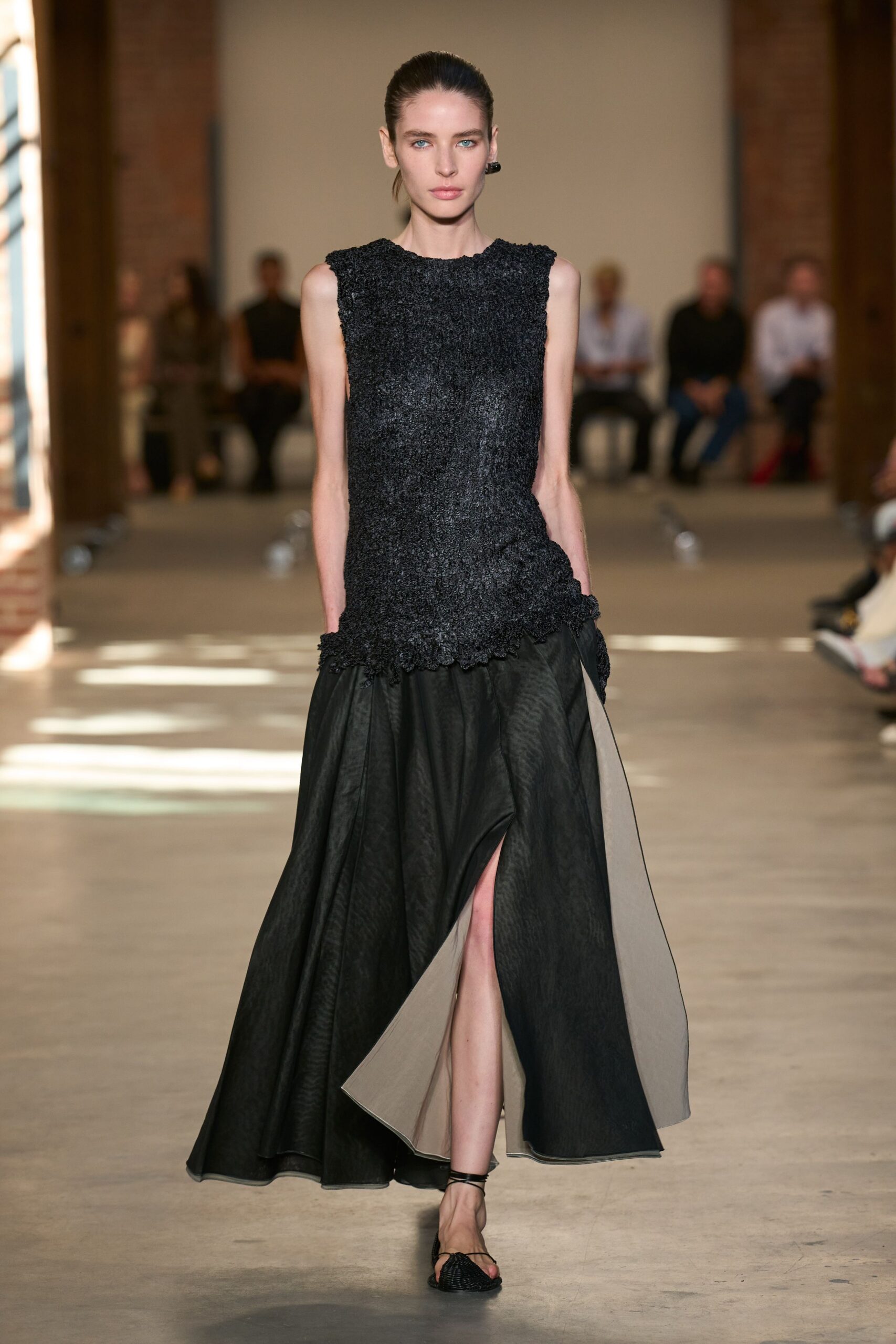 Charcoal boucle shell with raw scalloped edges and a slight peplum wobble. Black paper-taffeta ball skirt, split high to show its dove lining. Black thong sandals with a woven vamp. Single glossy black drop earring. Crossing a museum atrium while the afternoon sun moves across the floor.
