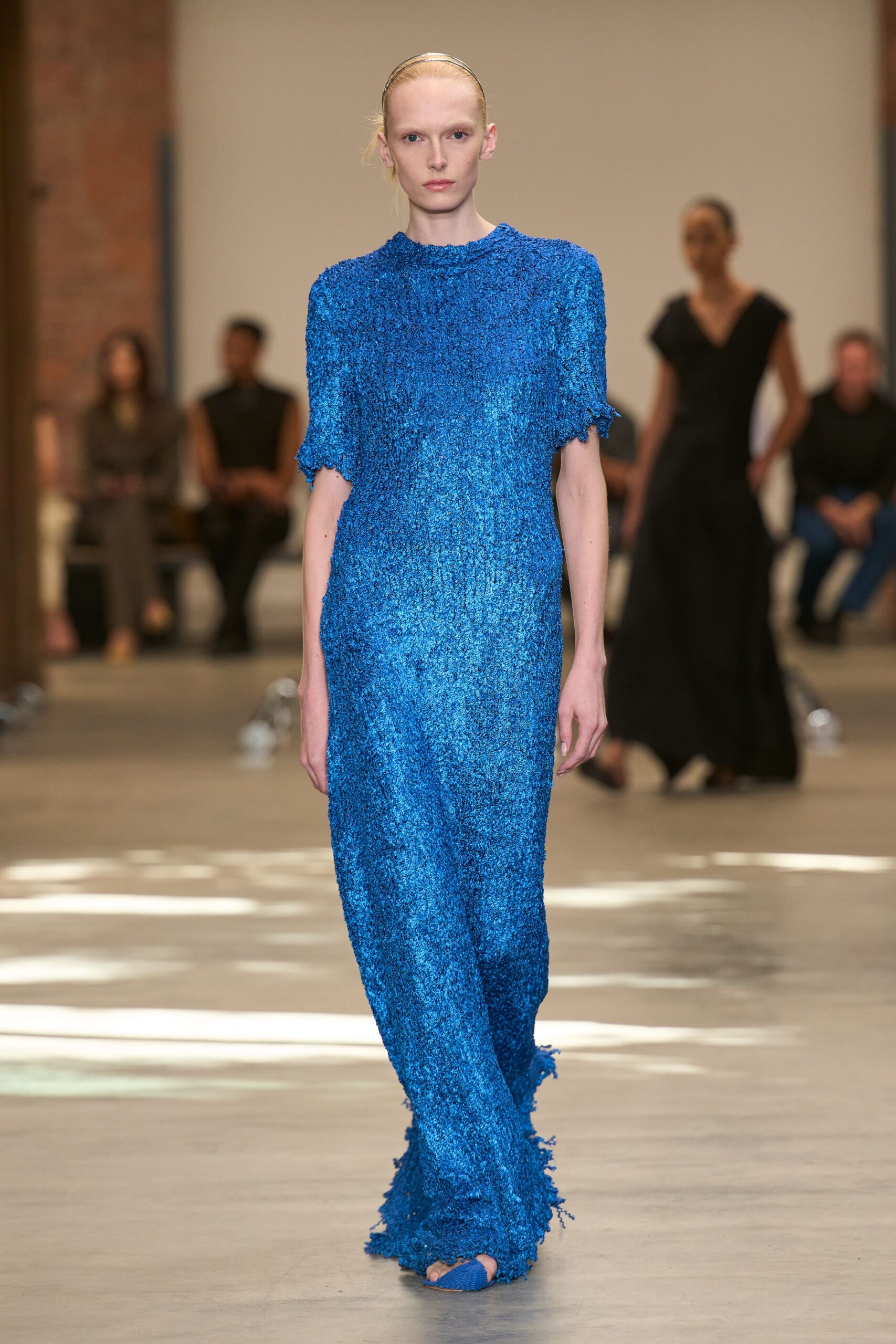 Slim metallic headband flattening the hair at the crown. Electric-blue bouclé column dress with ragged cuffs and a brushing hem. Blue woven peep-toes barely visible under the pooled edge. Crossing a museum atrium while the floor still holds afternoon heat.