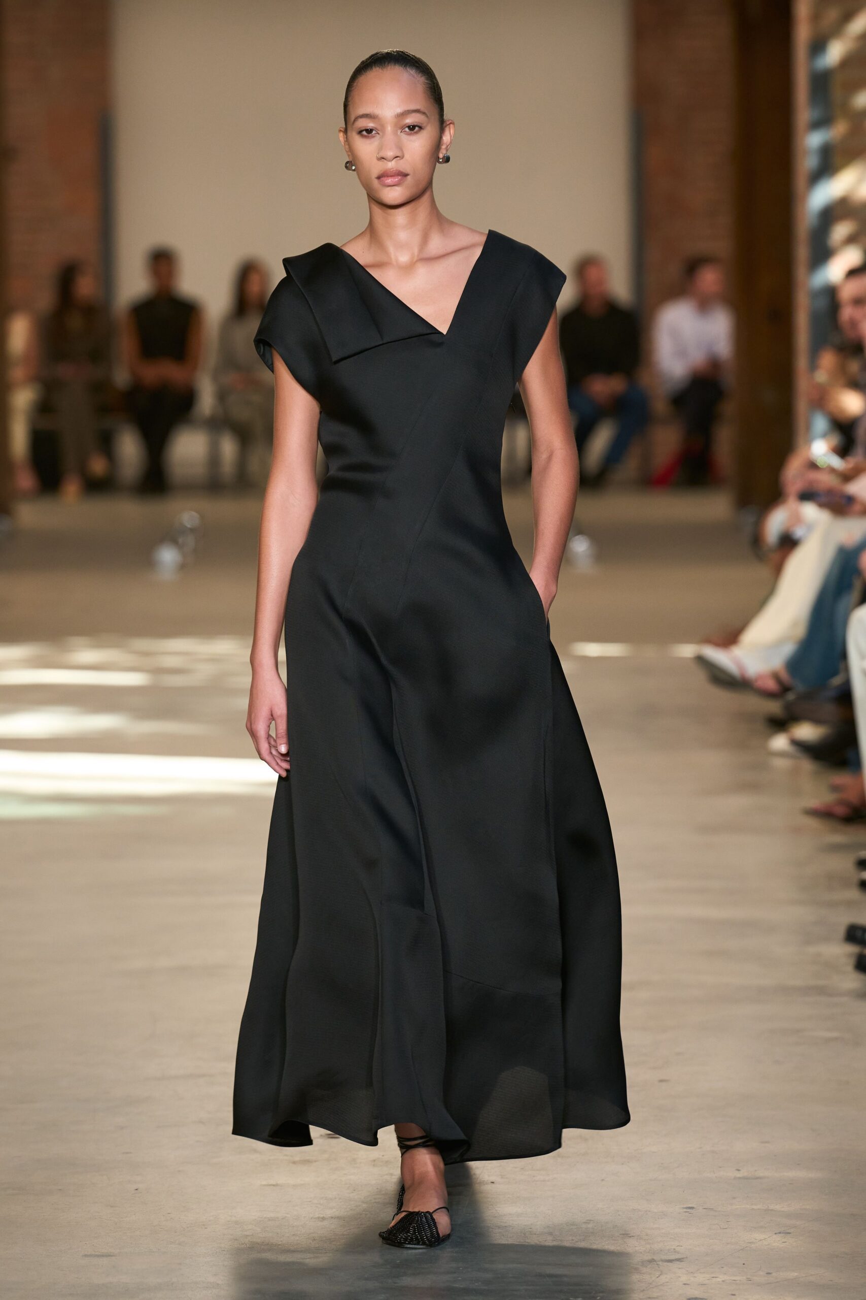 Small dark hoops hugging the lobe. Black crepe dress with a folded origami shoulder and slant seaming. Pointed mesh flats, ankle ties crossing sparely. Crossing a hushed museum atrium before the doors open.