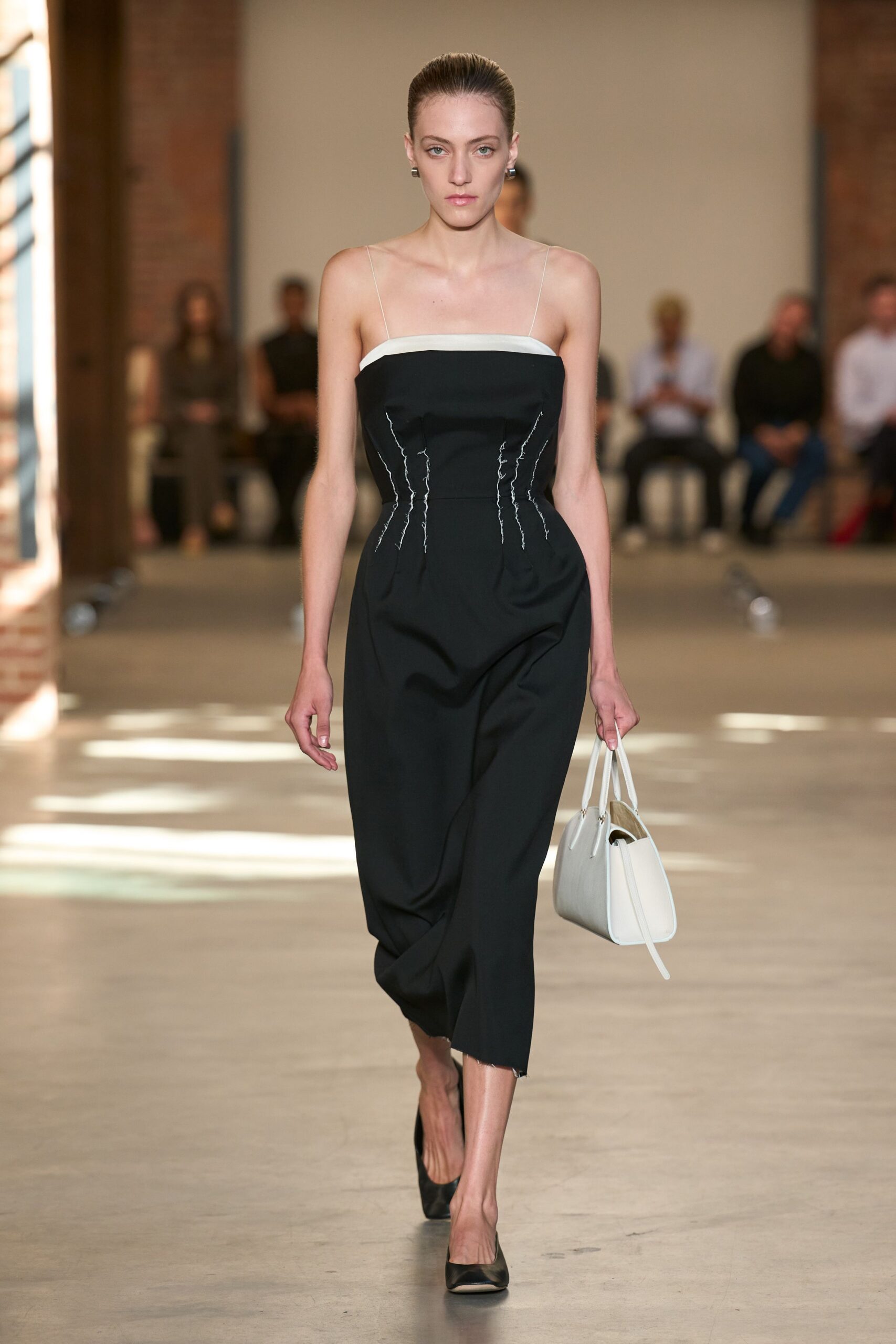 Black column dress with ivory binding and wiry seam traces cinching the torso. White trapeze tote, long strap trailing past the base. Black pumps with a low vamp and pointed toe. Small silver block earrings. Crossing a museum atrium before the gift shop opens.