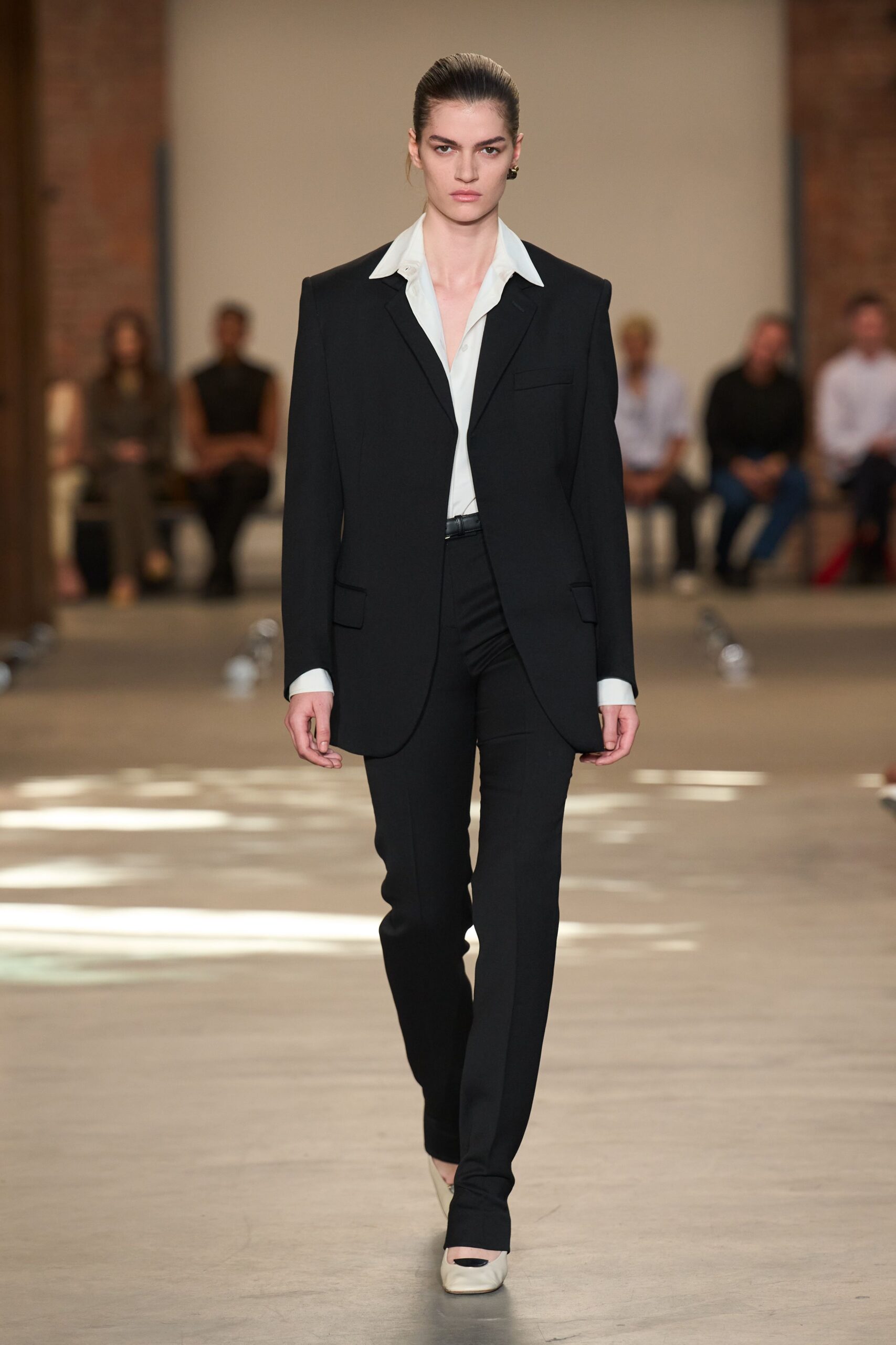 Black tailored blazer with a long curved front and flap pockets. White shirt, wide collar spread open and cuffs left long. Slim black trousers breaking slightly over the instep. Black leather belt with a small silver buckle. Cream pumps with a black cap toe. For the meeting after lunch when the hotel bar is still empty.