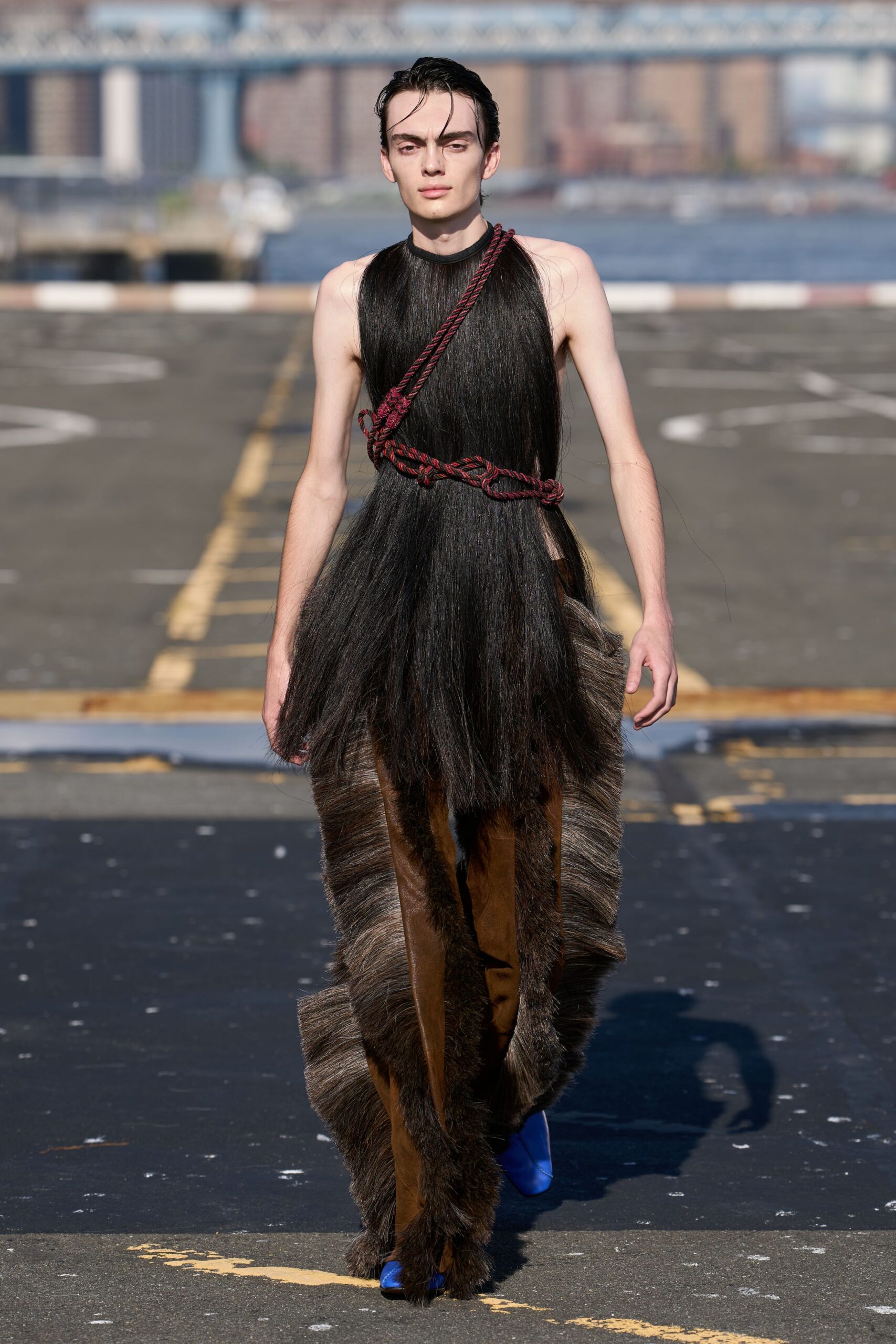 Black horsehair halter dress falling in glossy curtain lengths. Burgundy-and-black twisted rope knotted across the chest and waist. Brown leather trousers with shaggy grey fur panels swallowing the legs. Cobalt pointed boots flashing out beneath the furred hem. Crossing the ferry lot after rain, wind off the river still cold.