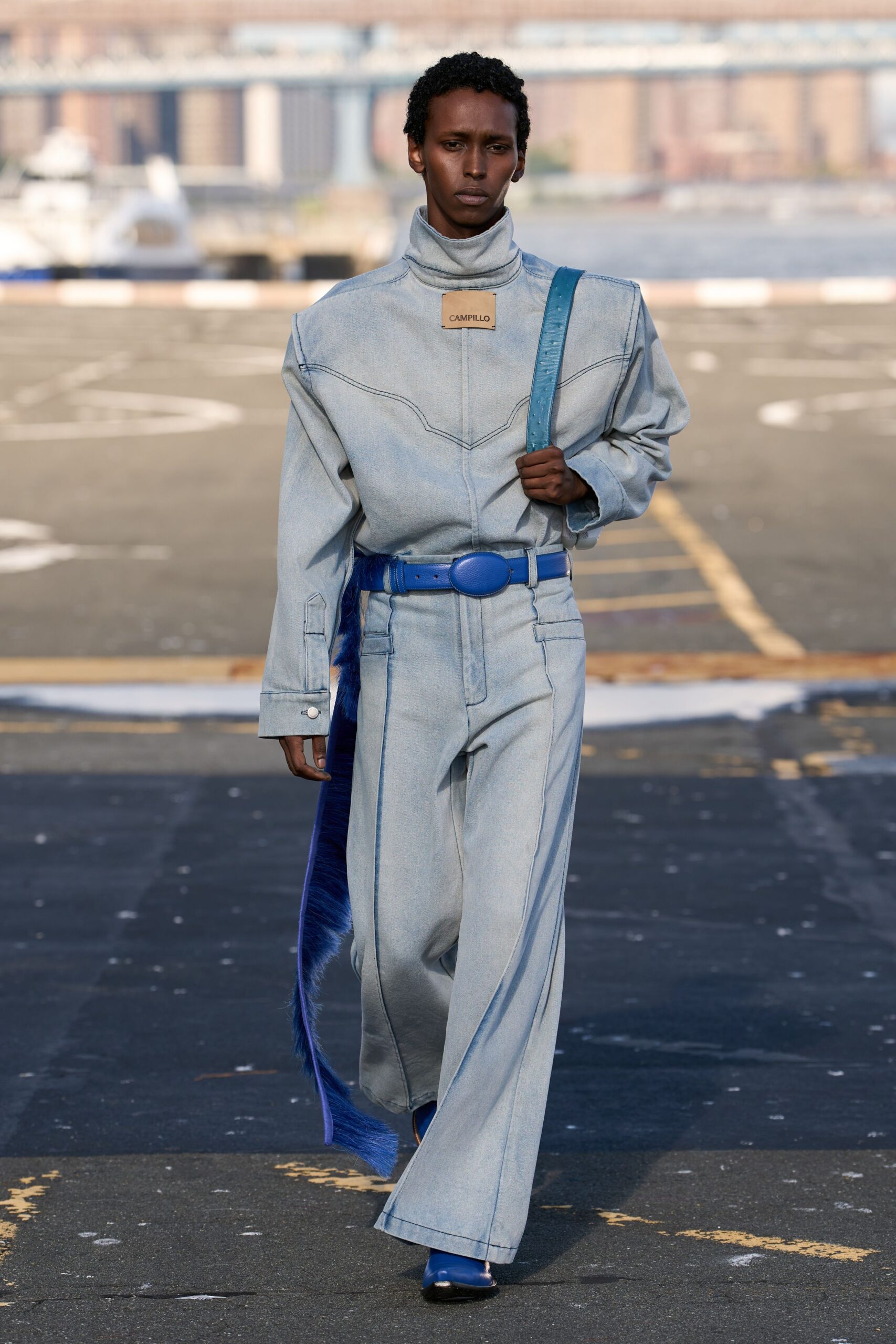 Pale stonewashed denim jumpsuit with a high rolled neck and curved yoke seaming. Cobalt leather belt with an oval buckle and long fur tail. Blue leather strap crossing one shoulder. Pointed cobalt boots peeking under the wide hem. Crossing the ferry lot before sunrise with salt drying on the railings.