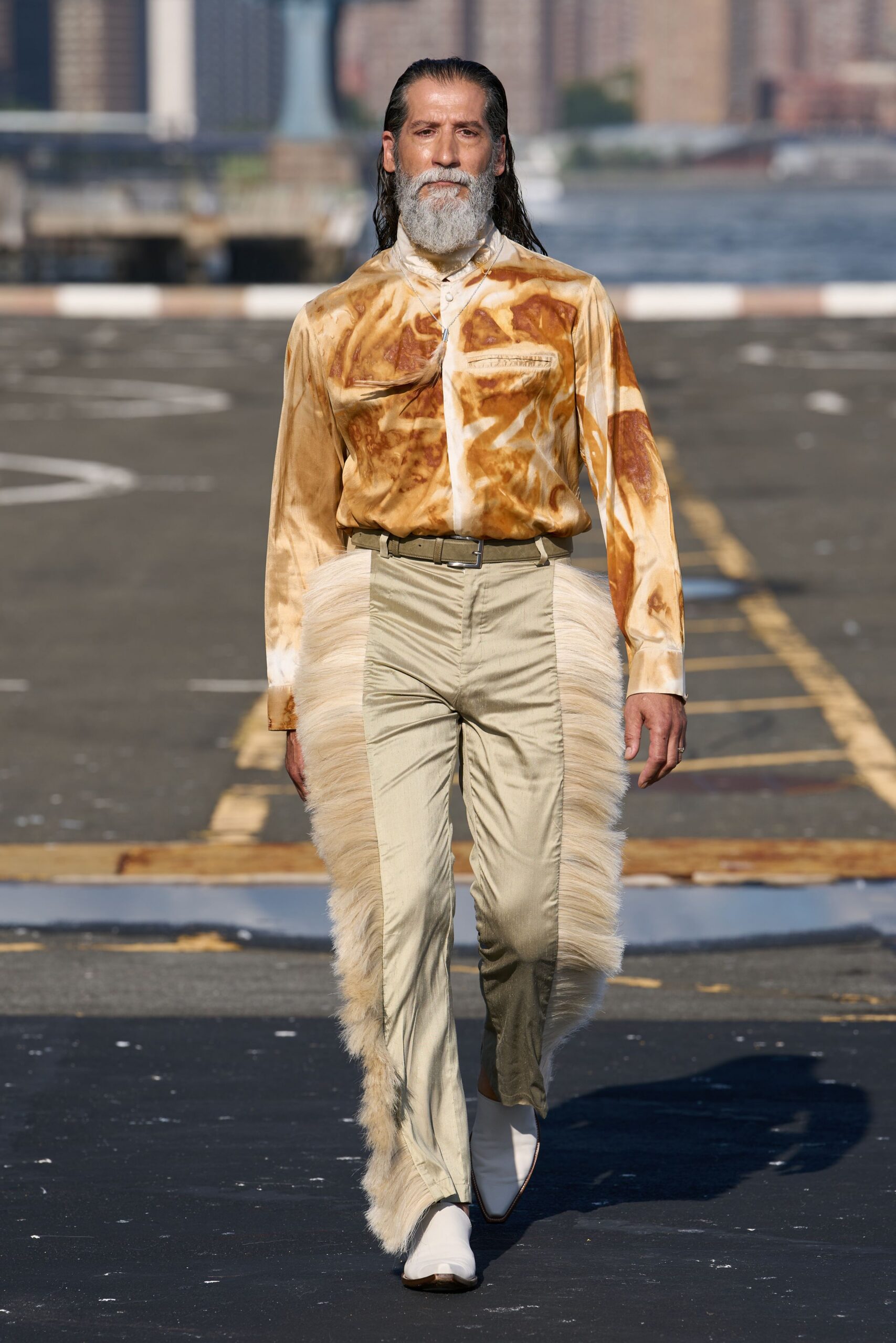 Glossy cream shirt marbled in toast-brown swirls, chest zip welt slightly puckered. Fine chain with a slim blue pendant. Olive web belt with a square metal buckle. Pearly narrow trousers flanked by shaggy cream horsehair panels. White square-toe ankle boots. Crossing the ferry lot after sunrise, still carrying hay on your cuffs.
