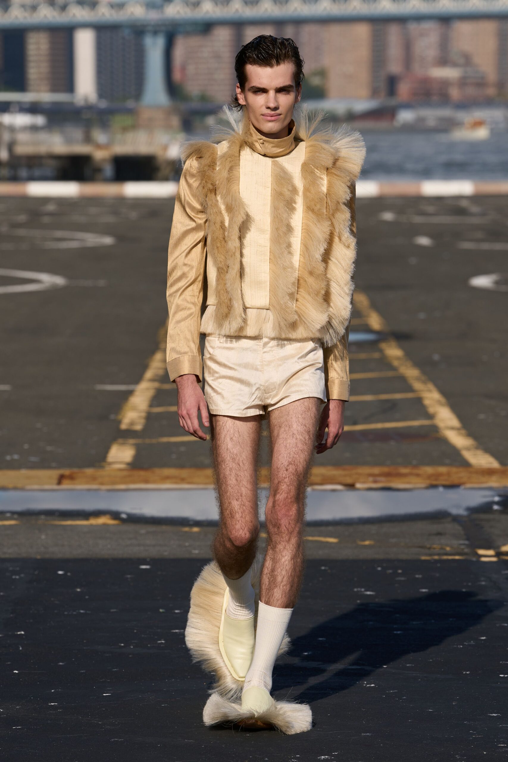 Champagne satin top with a high neck and long fur panels. Pearly silk shorts cut very short, creased across the fly. Cream ribbed tube socks. Pale slip-ons ringed with long cream fur at sole and toe. Crossing the ferry deck in cold sun, pretending bare legs make sense.