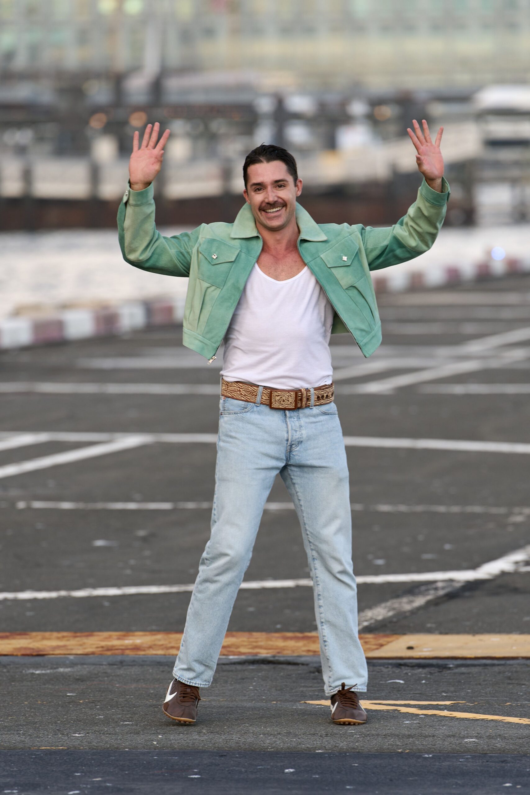 Mint suede jacket cropped short, silver snaps on the flap pockets. White jersey tank with a wide scooped neckline. Tooled tan leather belt with a large square buckle. Pale stonewashed jeans cut straight through the leg. Brown leather trainers with gum soles. Waving across the ferry lot before the last car boards.