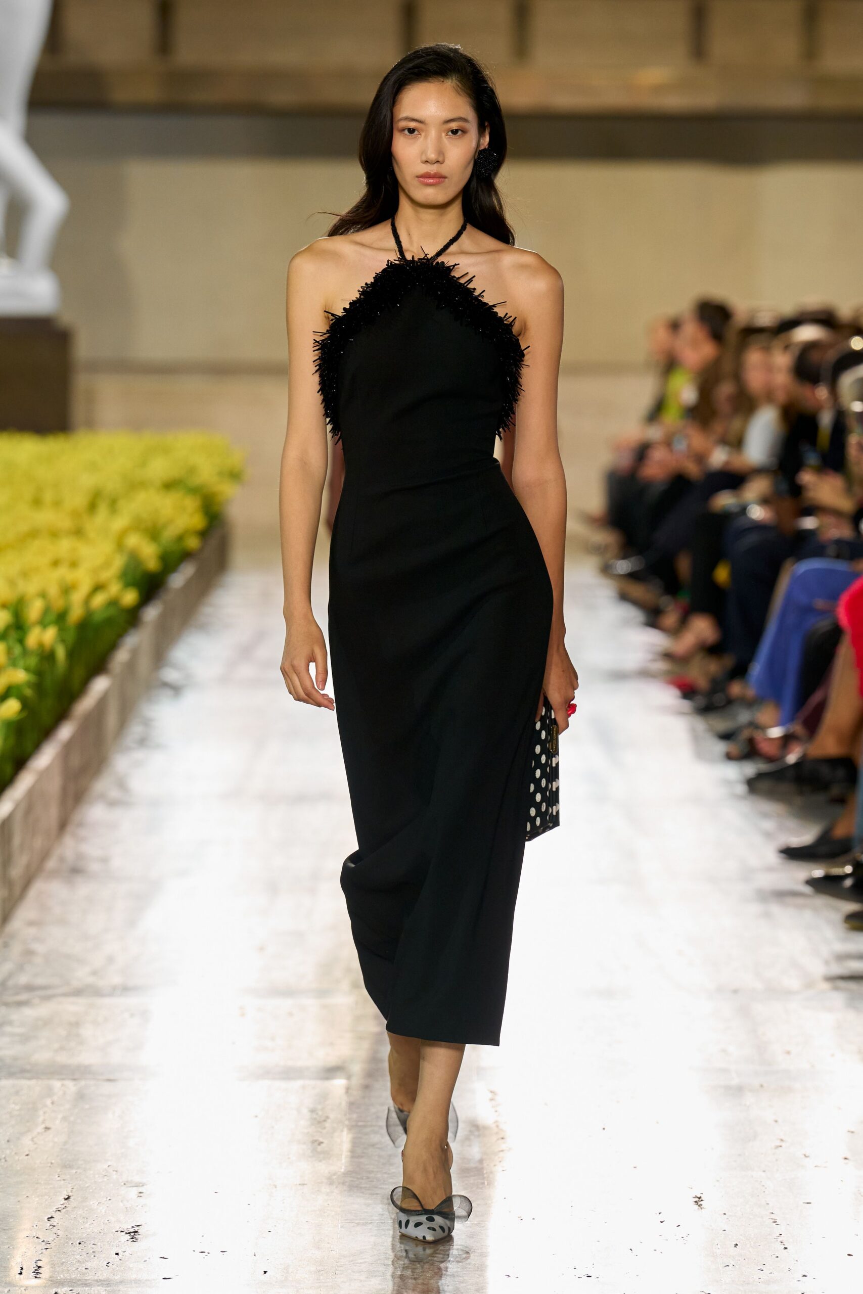 Black halter column dress with beaded fringe needling around the neckline. Single oversized black pom-pom stud. Box clutch in black satin, scattered with white polka dots. Sheer black mesh pumps with spotted pointed toes. Crossing a museum atrium while the florist is still replacing yesterday's tulips.