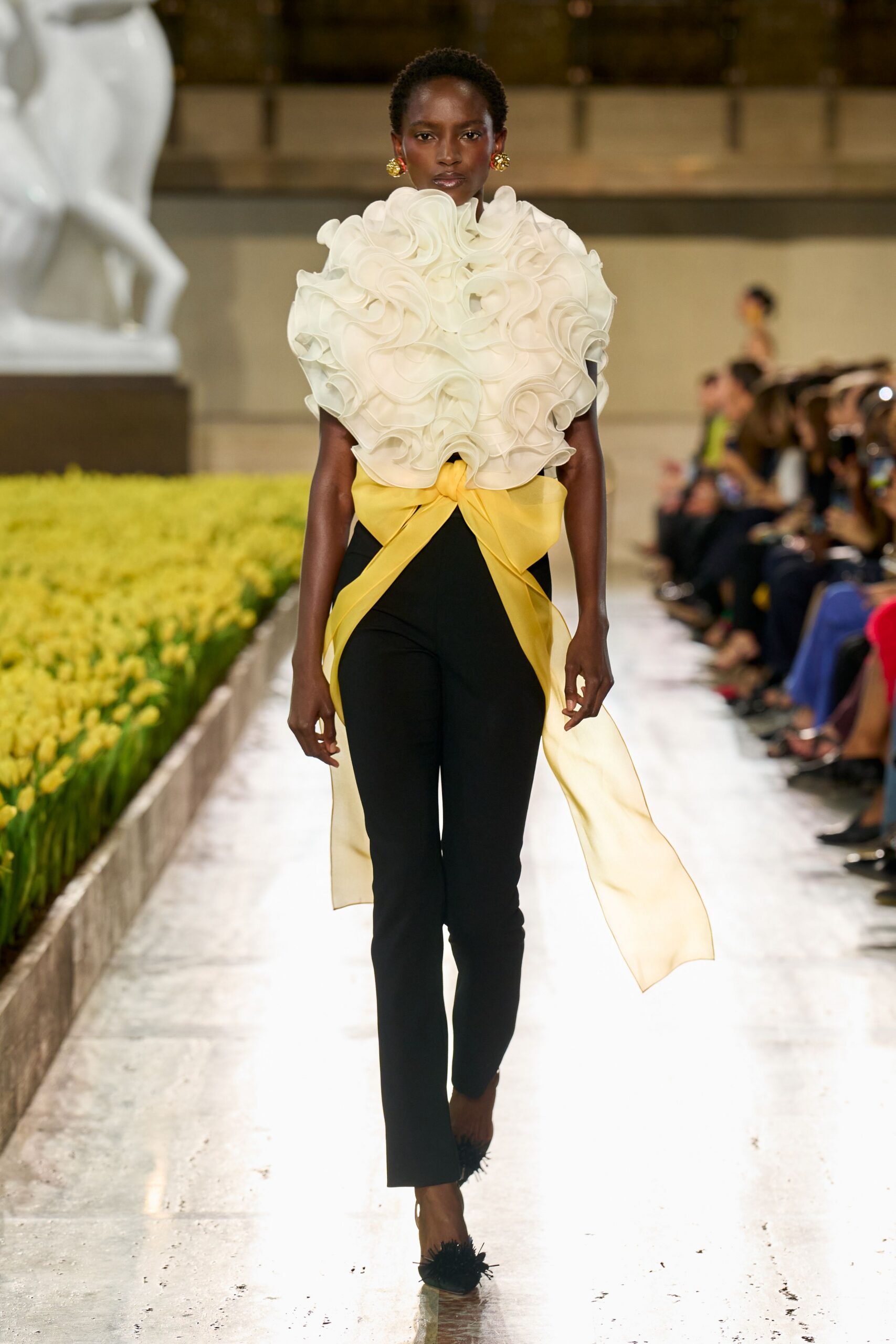 Ivory organza rosettes piled into a high sculpted bib. Butter-yellow bow tied low, one tail dragging nearly to the calf. Black slim trousers in clean matte crepe. Round floral clips in red and gold. Black flats with shaggy feathered toes. Walking the conservatory aisle while the tulips are forced early.
