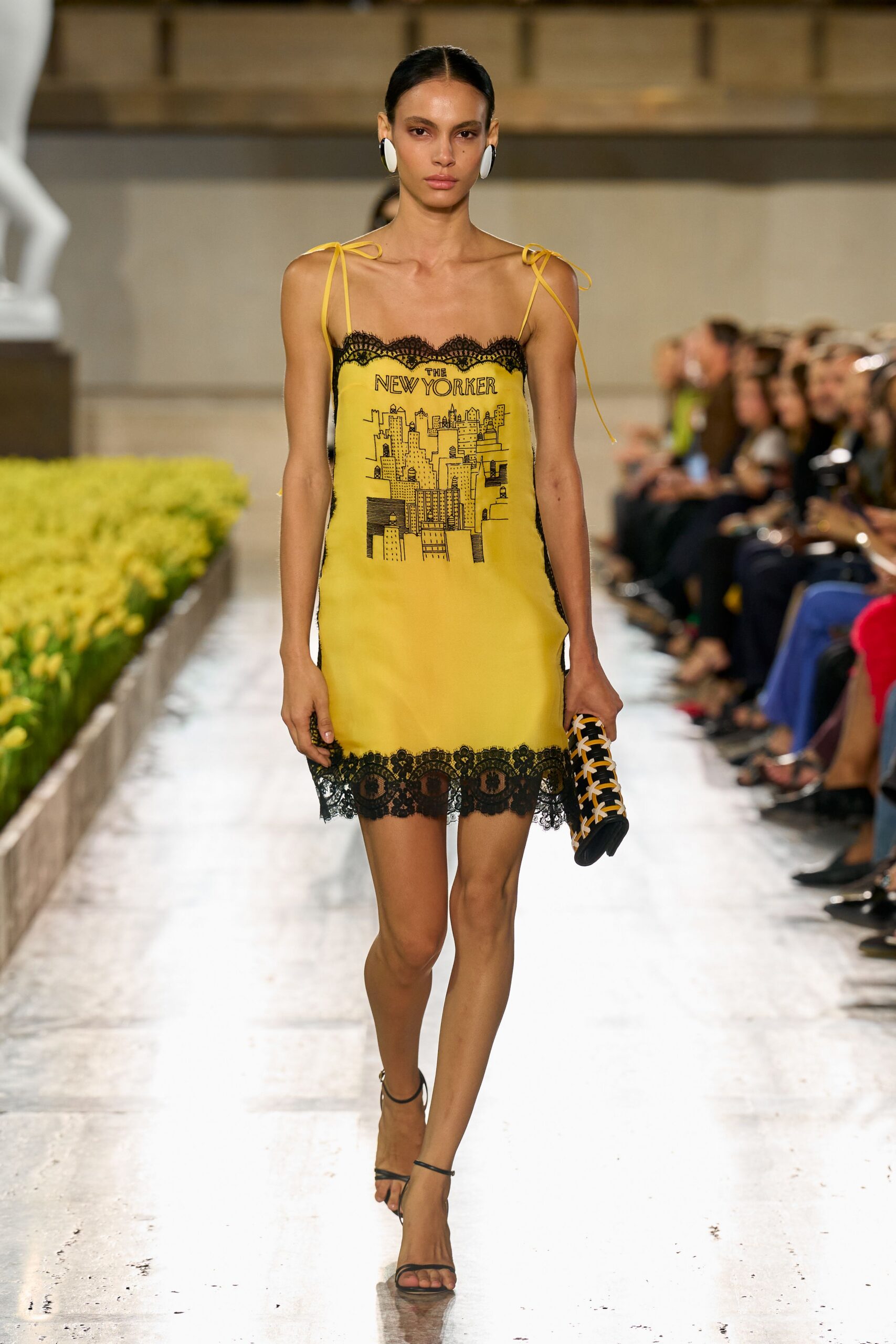 Canary slip dress with black lace edging and a hand-drawn skyline print. Oversized white oval discs. Black cylinder clutch latticed with mustard and white cords. Bare black ankle-strap sandals. Walking past florist buckets with tomorrow's tabloid tucked under your arm.
