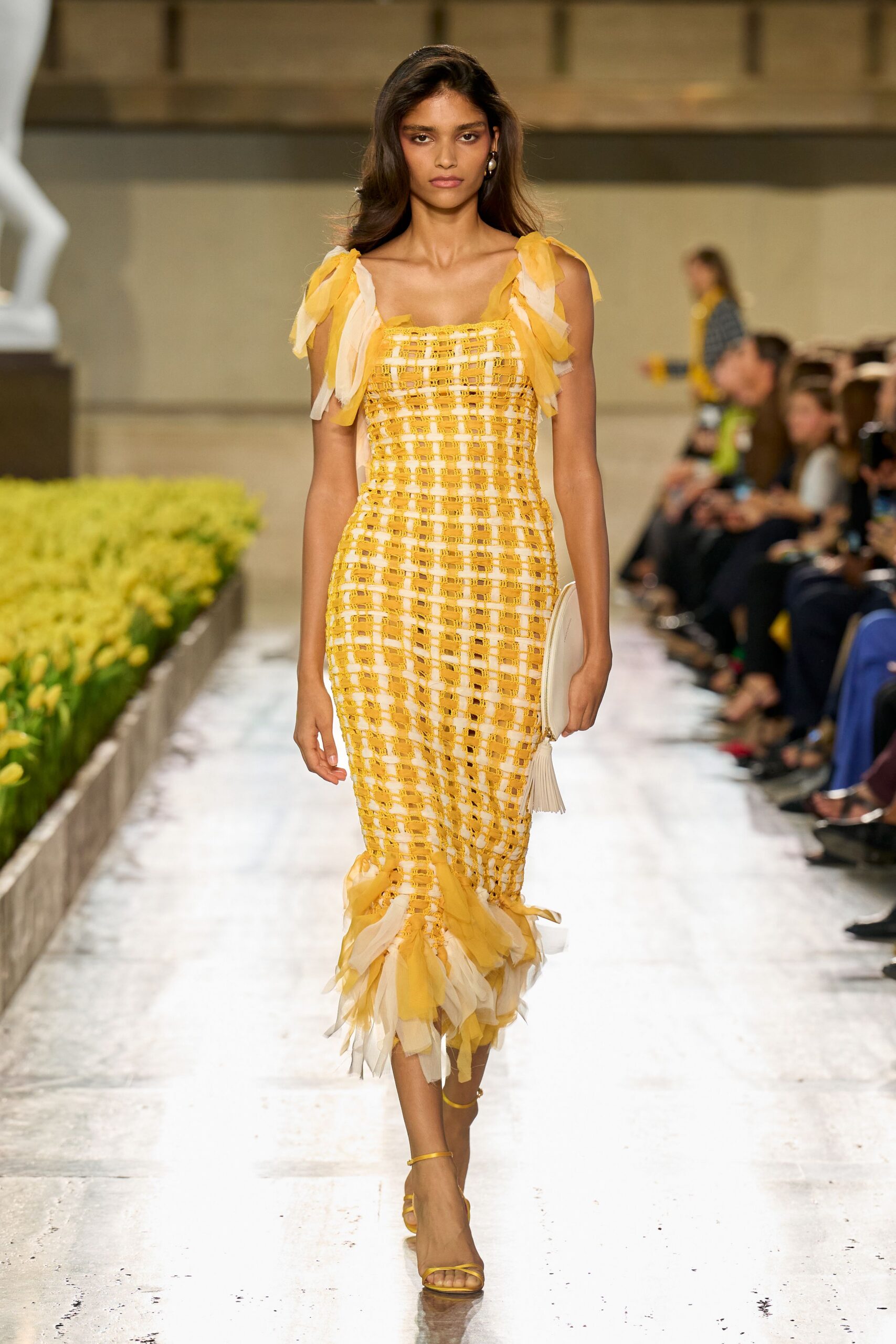 Butter-yellow openwork column dress over white lining, chiffon ties fluttering from straps and hem. Ivory zip pouch with a long tassel swinging at the wrist. Thin yellow ankle-strap sandals. Mismatched pearl drop earrings. Walking past greenhouse beds just after the first daffodils opened.