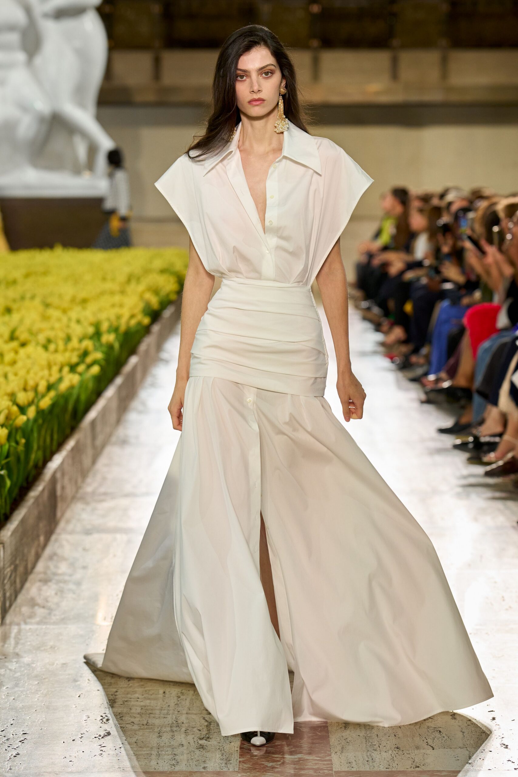 White cotton poplin shirt dress with a plunging placket and wide cap sleeves. Broad self-fabric bandage wrap ruched low around the hips. Mismatched gold chandelier drops brushing the shoulder. Black pointed boots with white cap toes peeking out. Crossing a museum courtyard after the wedding, tulips still holding the day's heat.