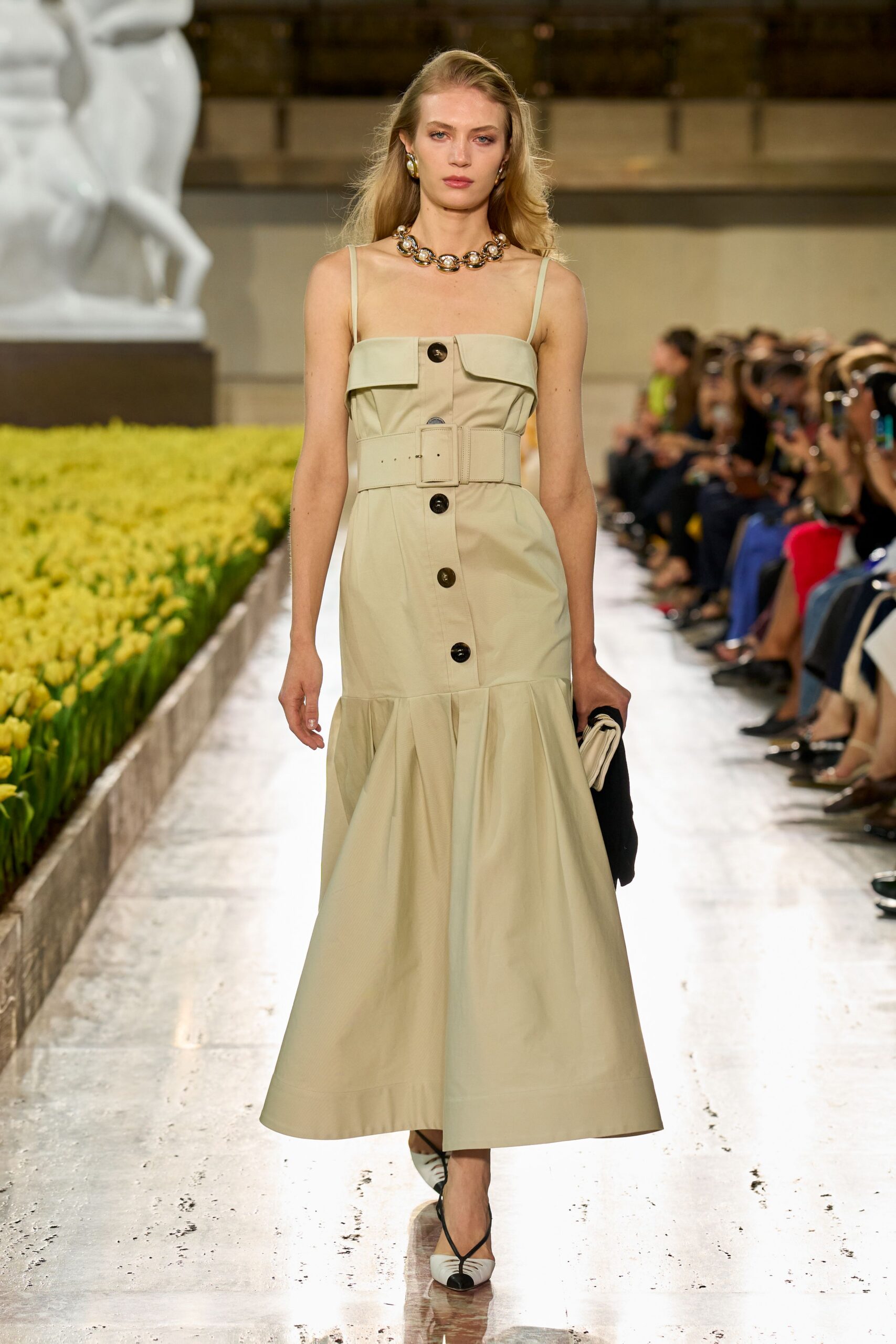 Pale sand cotton dress with a folded bib, horn-look buttons and wide belt. Oversized linked stones in a heavy collar. Matching oval drop earrings. Cream folded clutch tucked under a black layer. Cream slingbacks with black cap toes and crossed straps. Walking past formal beds of tulips before the museum opens.