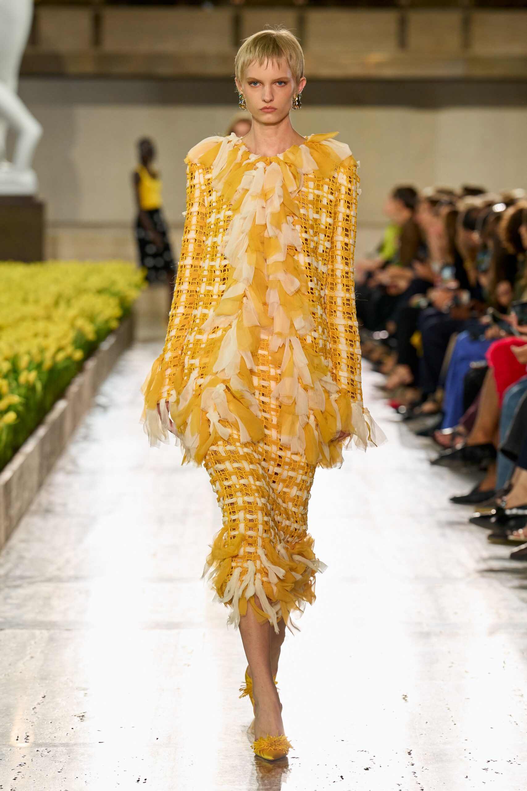 Marigold open-lattice column dress strewn with ivory chiffon ruffles down the front. Clustered jewel drops at each ear. Yellow satin pumps with frayed tassels at the pointed toe. Crossing a conservatory aisle while the last daffodils are being cut.