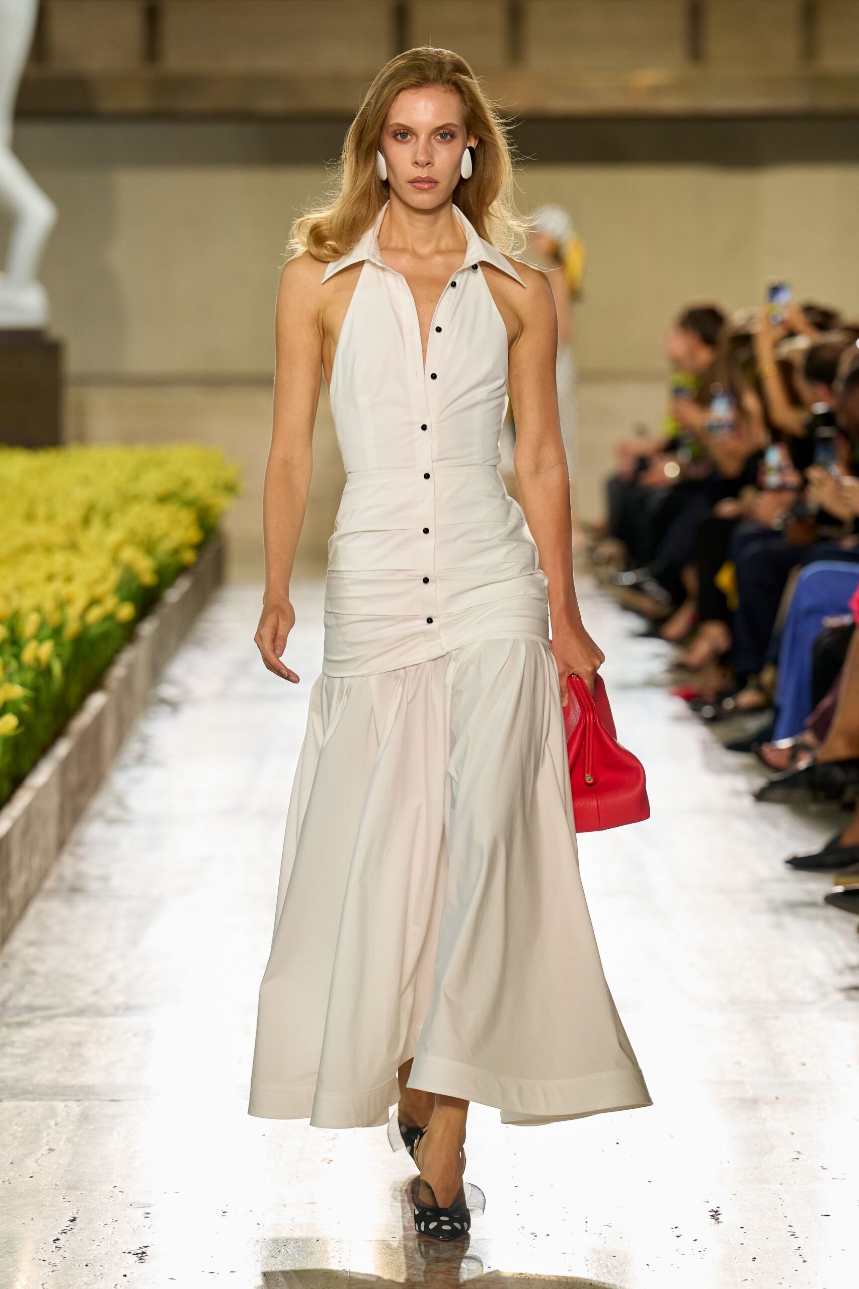 Large white oval clips at the lobes. Ivory halter shirt dress, black buttons and ruched hips above a dropped flounce. Tomato red leather hobo tucked under one hand. Black mesh slingbacks with pale spots at the pointed toe. Crossing a museum courtyard after the wedding lunch ran long.