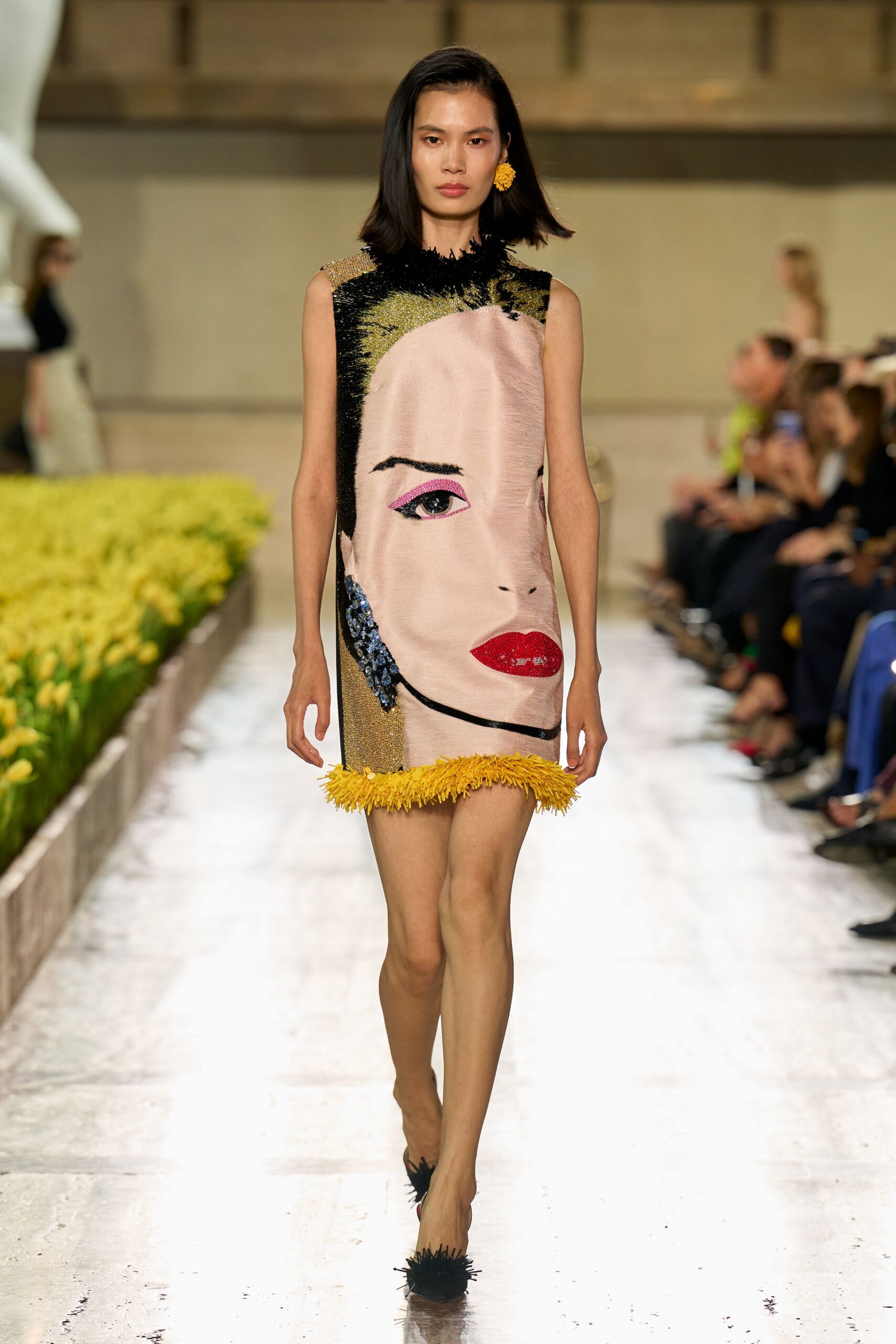 Sleeveless shift woven into a giant face, yellow fringe at hem and collar. Single marigold cluster earring. Pointed pumps with black raffia tufts at the toe. Walking past conservatory flowerbeds after noon, still carrying lipstick from breakfast.