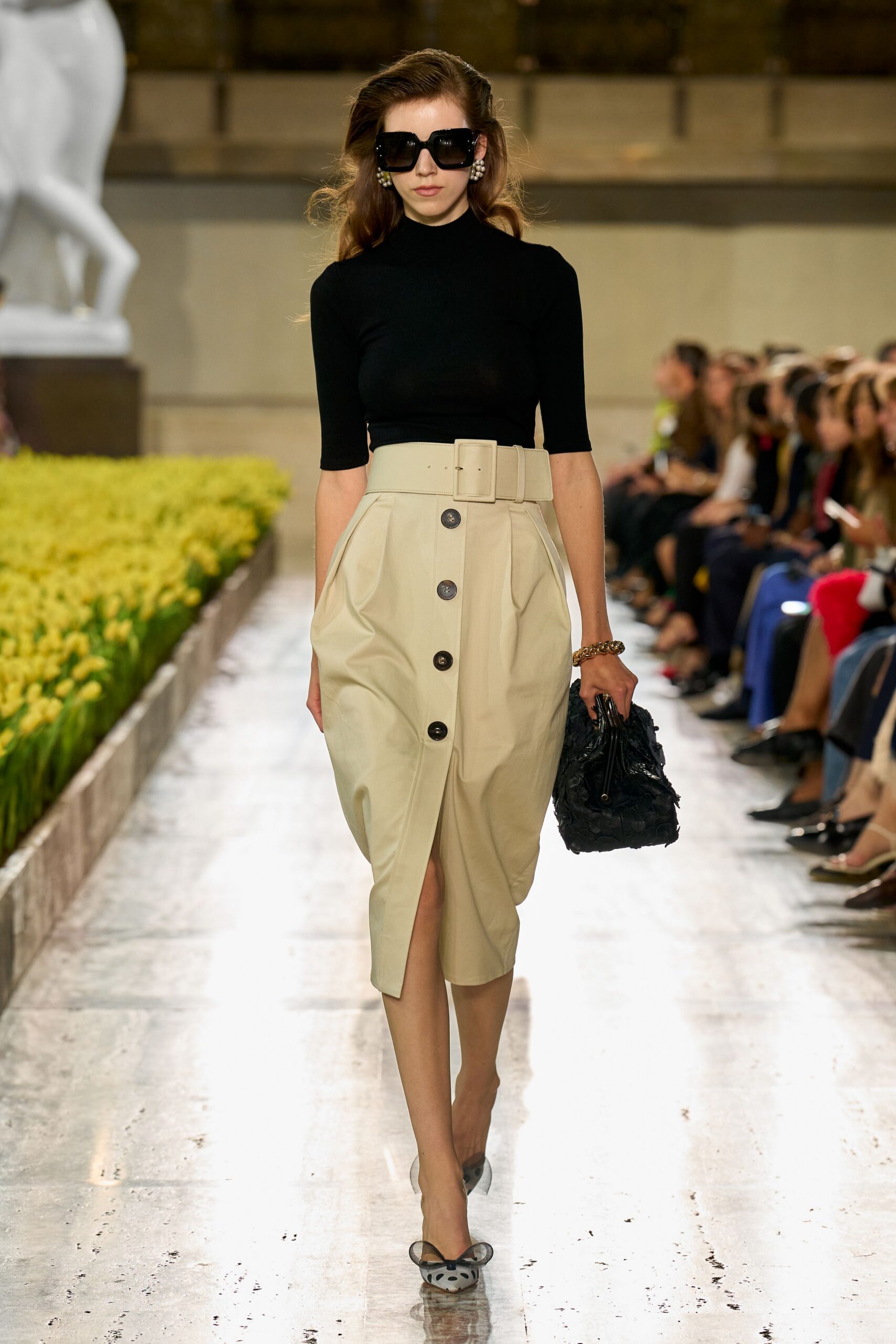 Oversized square black frames. Black knit mock-neck top with close half sleeves. Cream wrap skirt, paperbag waist cinched by a broad covered belt. Black rosette-textured satchel with short handles. Sheer mesh pumps, bow-tipped and dotted at the toe. Walking beside formal flowerbeds while the museum is still closed.