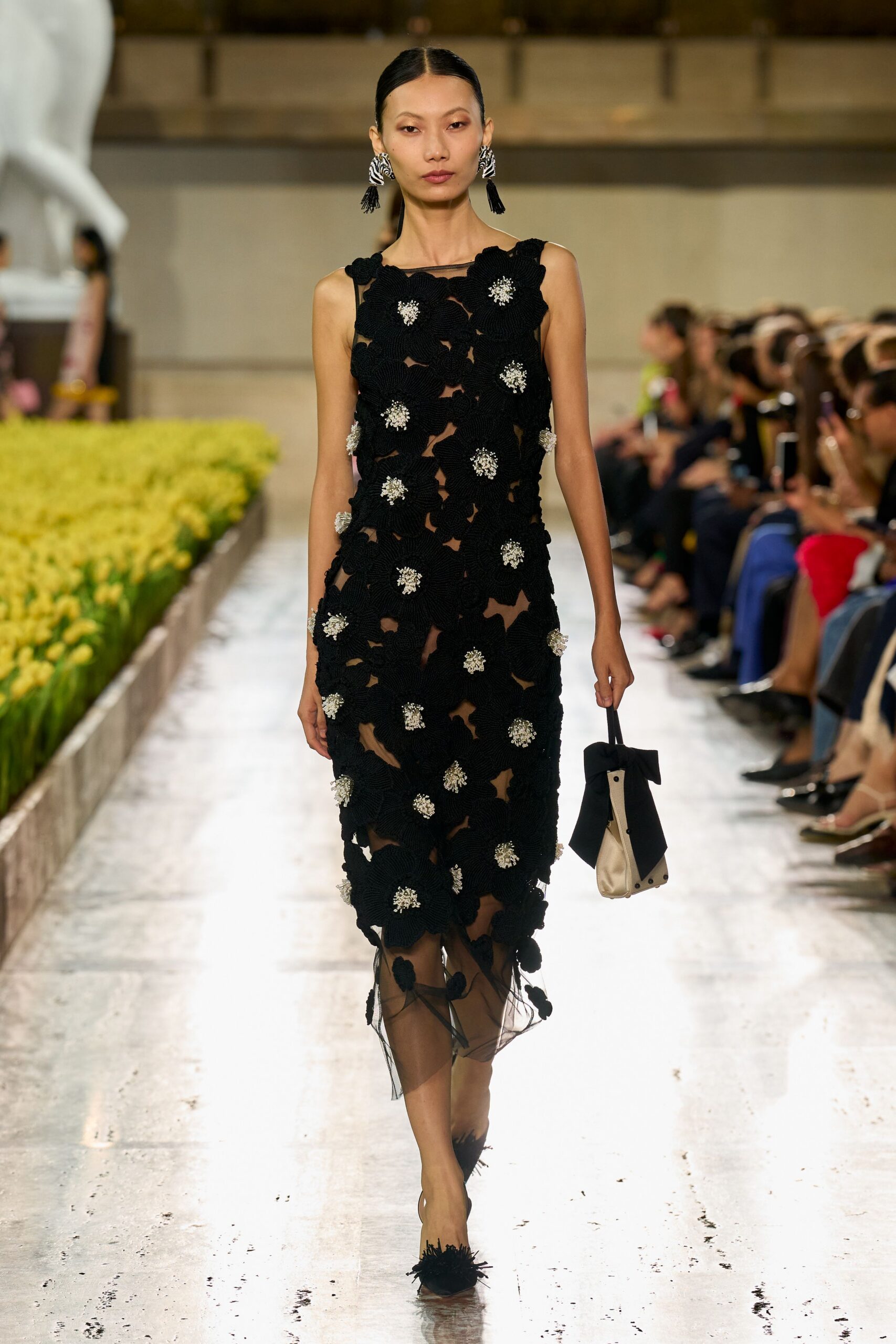Black floral crochet column over sheer mesh, crystal-cluster centers catching at each bloom. Zebra-striped tassel drops. Cream satin mini bag pinched under an outsized black bow. Pointed mesh flats with black fringe pompons. Crossing a museum courtyard after dark while the fountain is still on.