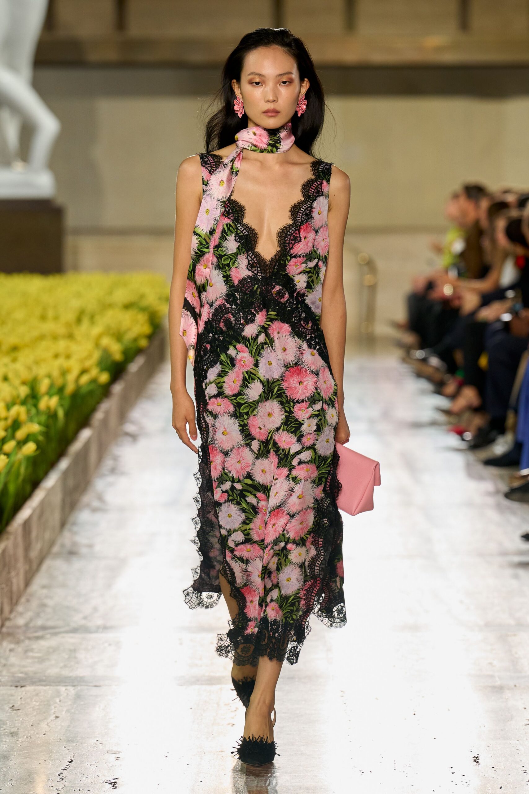 Pink flower cluster earrings. Silk neck scarf in the same pink chrysanthemum print. Bias-cut floral slip dress with black eyelash lace plunging to the waist. Folded bubblegum leather clutch under one arm. Black mules topped with shaggy fringe. Walking past greenhouse beds just watered, still dressed for champagne.
