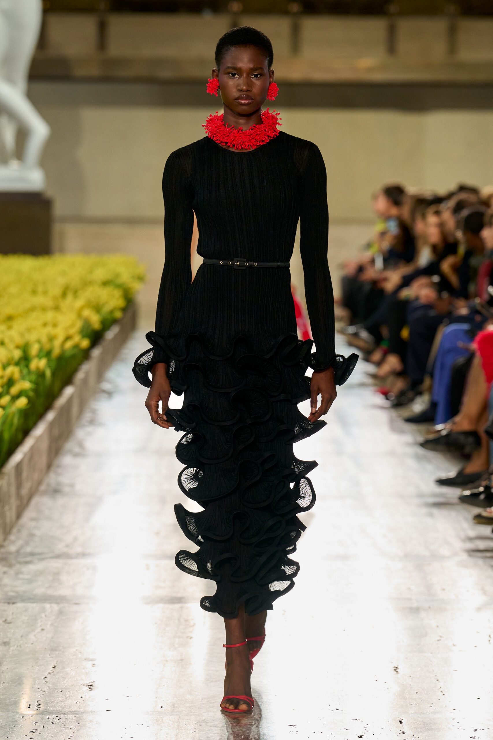 Bright red coral-like collar in dense branching clusters. Matching red cluster studs. Black ribbed knit column dress, hem built from looping wired ruffles. Narrow black belt with tiny silver eyelets. Red ankle-strap sandals. Crossing a museum atrium while the flower installation is still being watered.