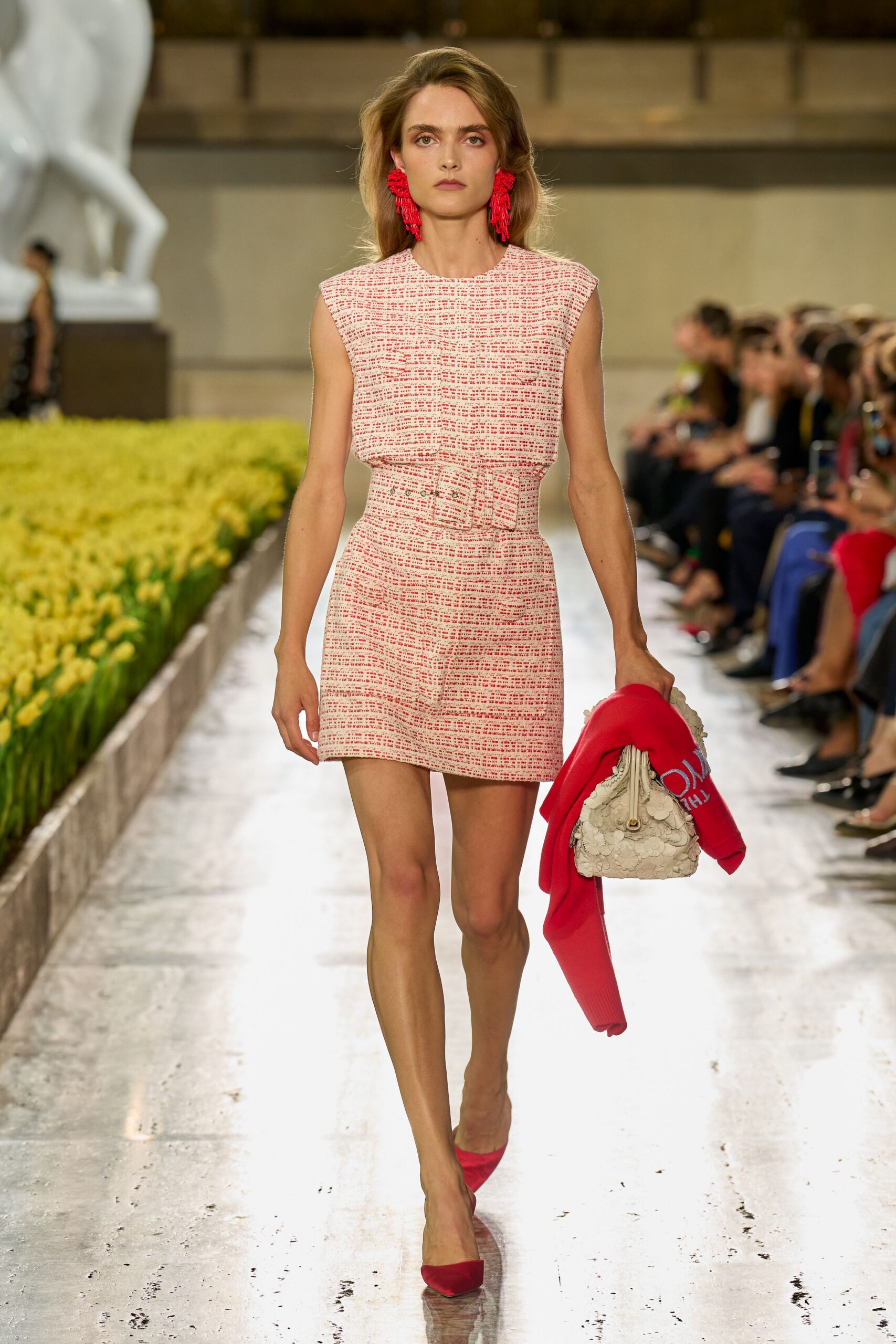 Coral beaded tassel earrings brushing the jawline. Pink and cream bouclé mini dress with a wide self belt. Cream bag covered in stitched petal appliqué. Hot-pink knit carried over one arm, white letters at the cuff. Pointed coral pumps in matte suede. Crossing a museum courtyard just after the flower beds were watered.