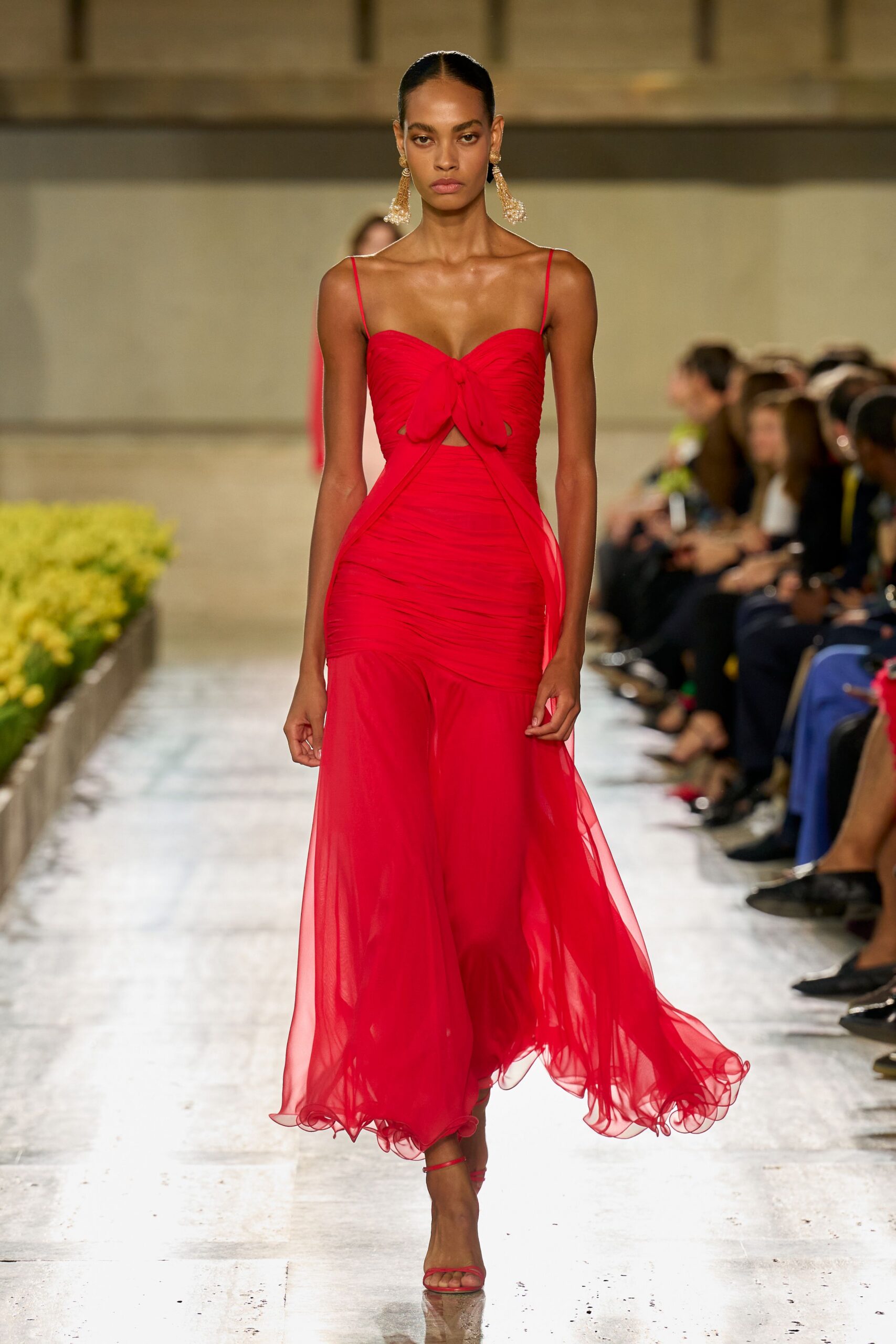 Long pearl tassel drops with small gold rosettes at the lobe. Scarlet chiffon gown, ruched through the body with a bowed bust cutout. Bare red ankle-strap sandals. For crossing a hotel courtyard just after midnight, heat still rising off the stone.
