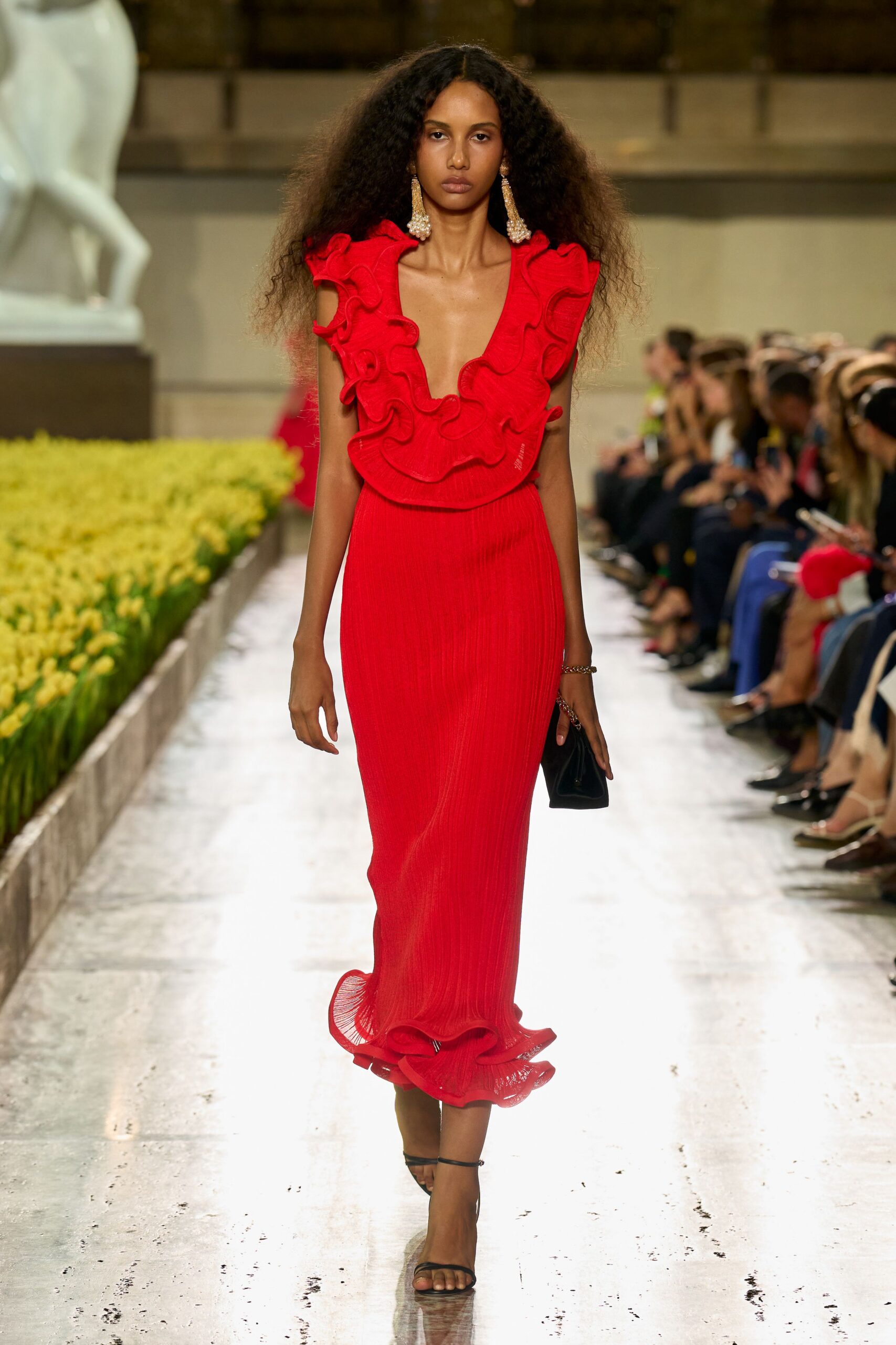 Scarlet pleated gown with a plunging V and rippling ruffle collar. Long pearl cluster drops brushing the shoulders. Silver chain bracelet at one wrist. Small black pouch in the hand. Black ankle-strap sandals, barely there. Crossing a museum courtyard while the fountain throws mist onto the stone.