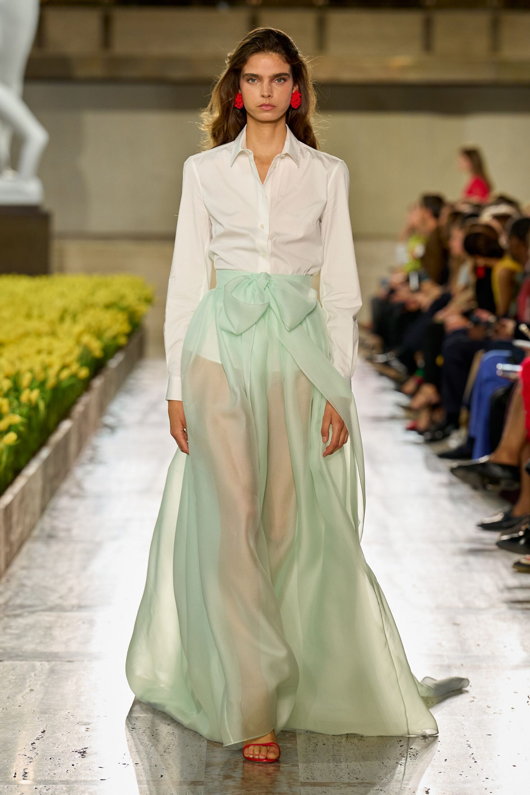 White cotton shirt, sharp collar and small mother-of-pearl buttons. Mint chiffon overskirt tied in a huge bow at the waist. Cream straight trousers visible beneath the sheer layer. Red barely-there sandals. Clustered scarlet earrings. Crossing a hotel courtyard just after the fountains switch on.