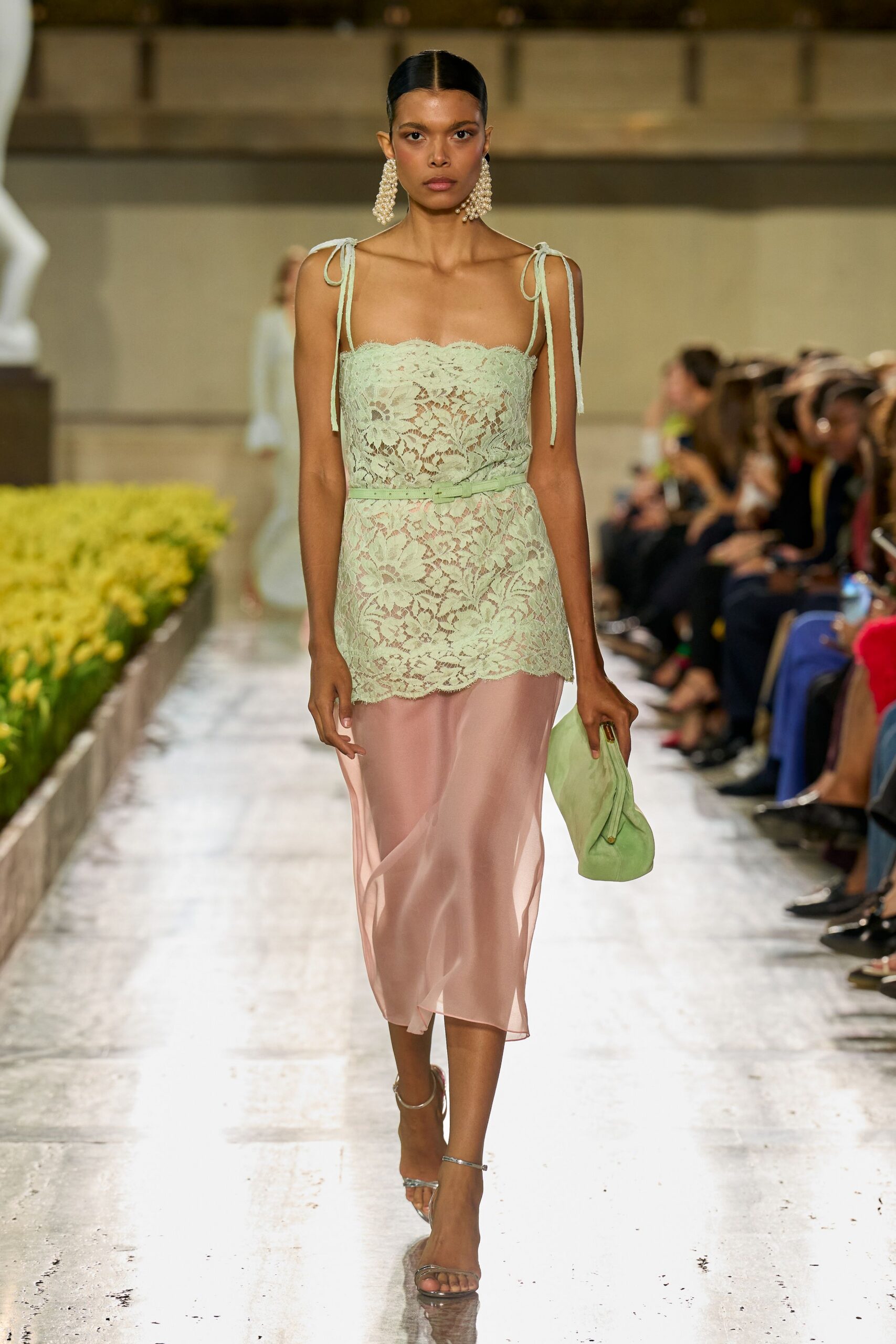 Long clustered pearl drops brushing the jaw. Mint floral lace bodice over a blush chiffon skirt, shoulder ties hanging loose. Skinny mint belt knotted flat at the waist. Soft pistachio suede clutch, folded under the hand. Bare silver ankle-strap sandals. Walking back from a garden wedding with grass still stuck to one heel.