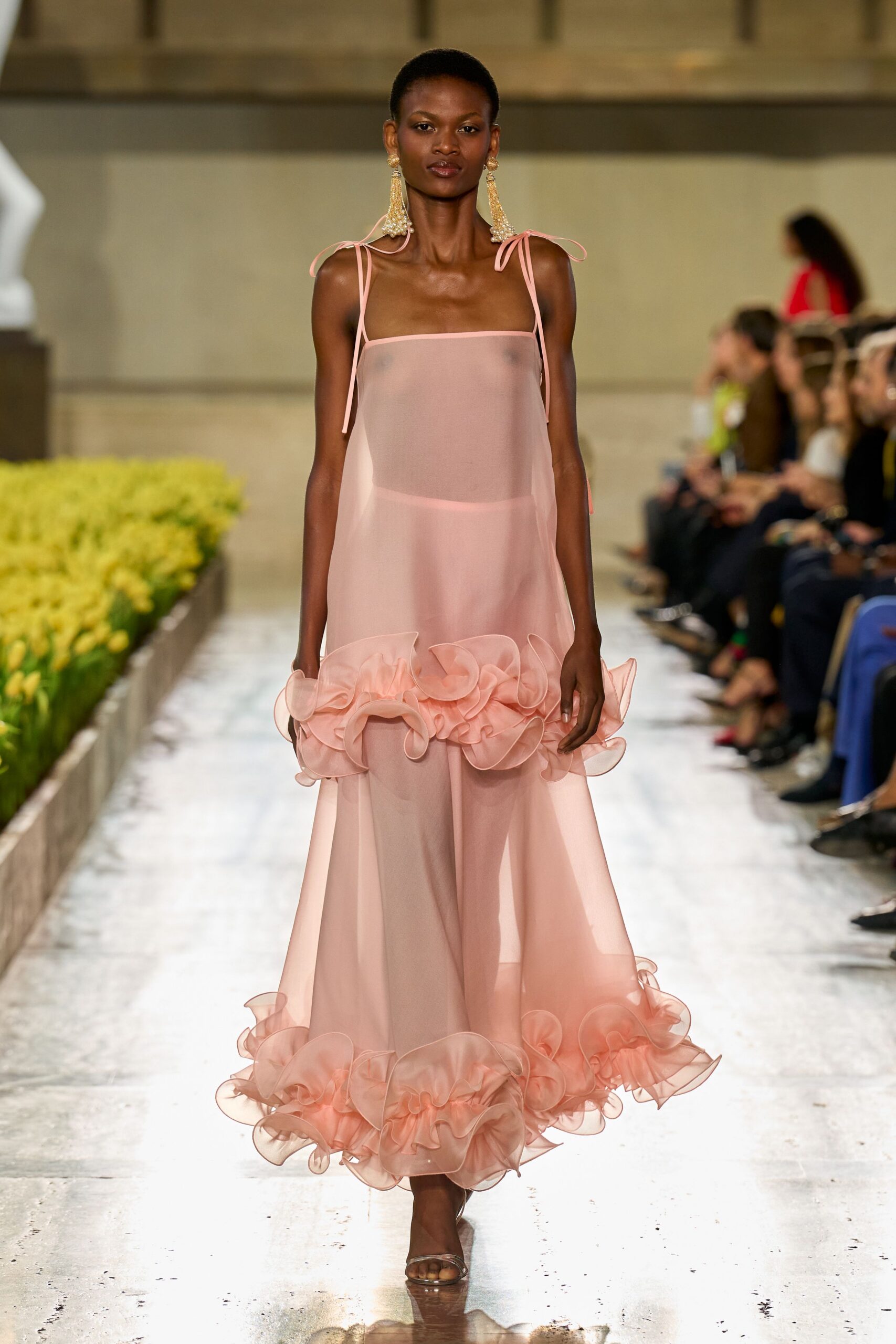 Long pearl-fringed earrings with a gold shell-like stud. Blush sheer organza slip dress, ribbon straps tied loose at the shoulders. Matching floor-length skirt, wired flounces curling at knee and hem. Clear strap sandals. Walking beside a conservatory pool while the orchid house fogs up.