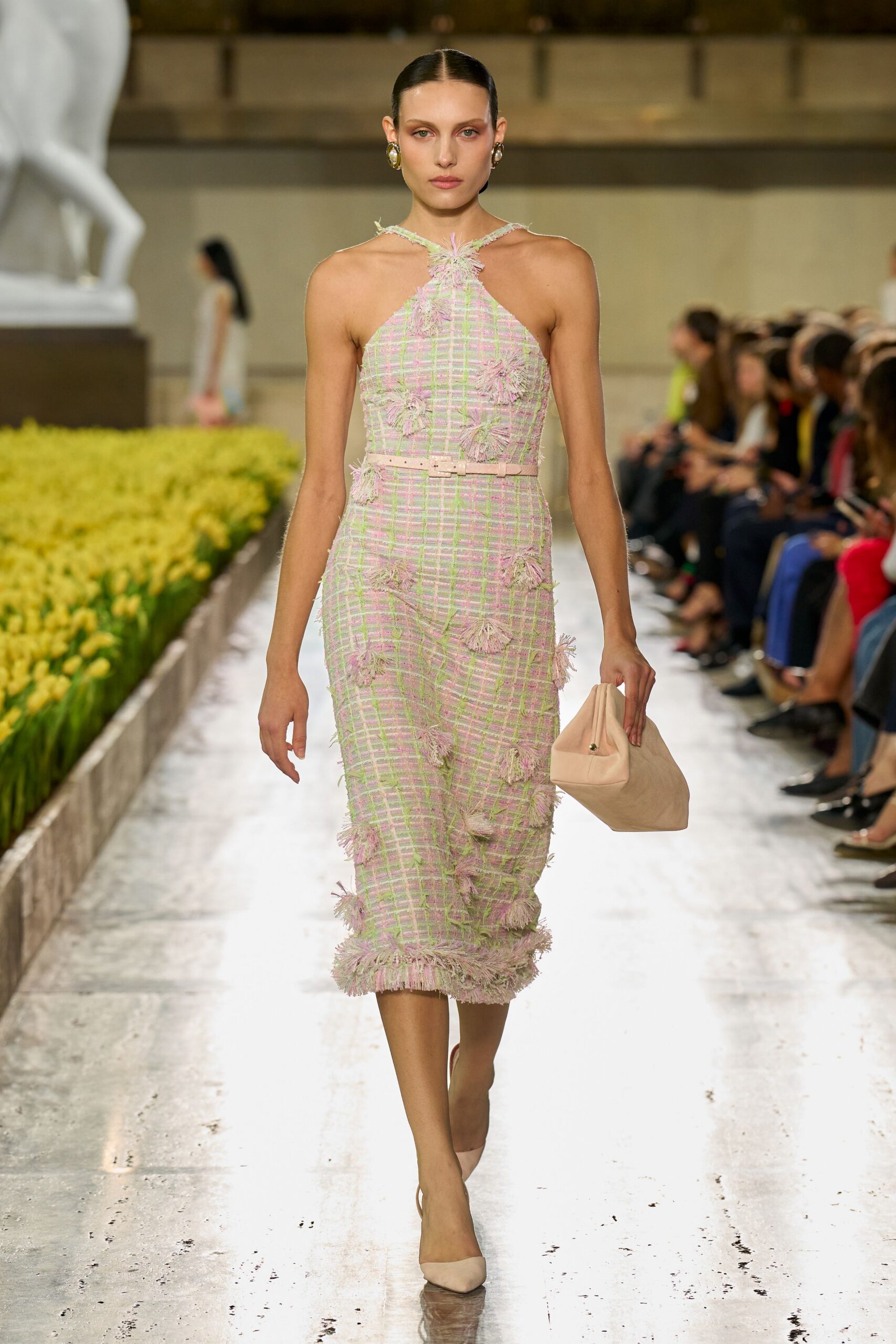 Pastel boucle halter sheath with tassel tufts and a narrow blush belt. Gold oval clips. Soft blush pouch, pinched shut at one corner. Nude suede pumps. Crossing a museum atrium while the florist is still replacing the tulips.