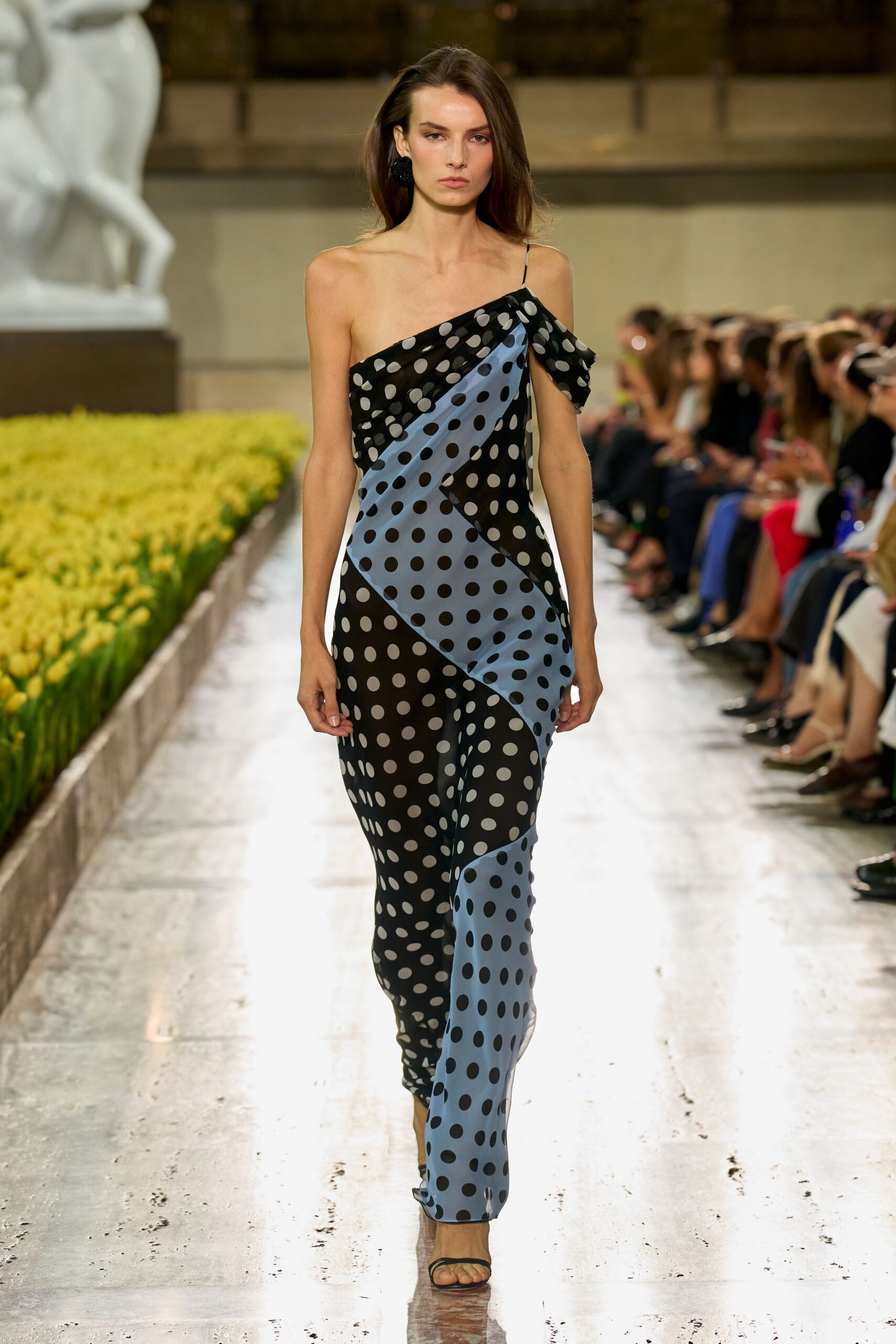 One-shoulder draped gown in black and powder-blue polka-dot chiffon, scarf panel falling from the strap. Oversized black rosettes at the lobes. Barely there black thong sandals. Walking past formal flower beds after rain, invitation folded in one hand.