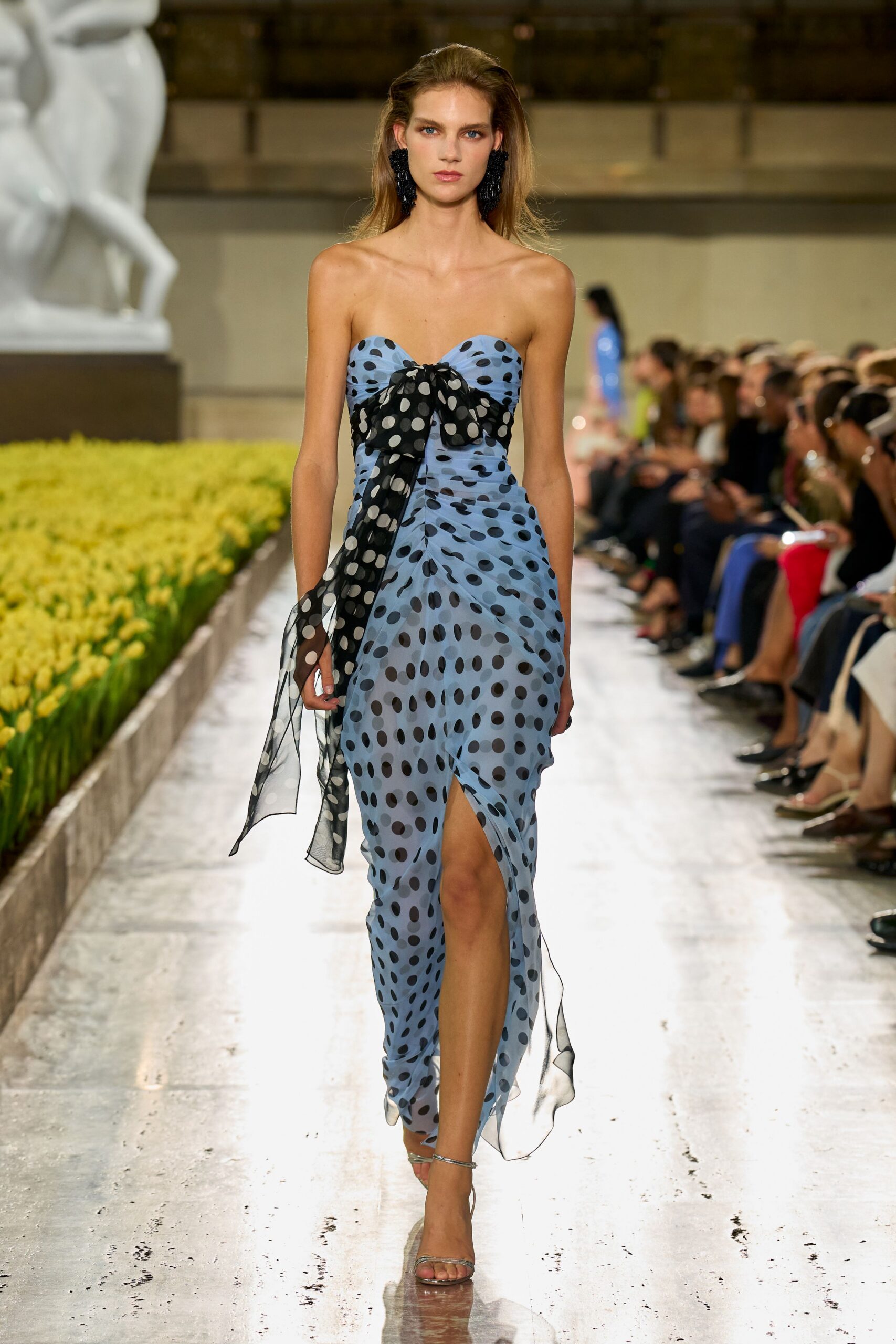 Black beaded tassel earrings brushing the jaw. Powder-blue strapless chiffon dress in black flocked dots, knotted with a long sheer bow. Barely there silver ankle-strap sandals. Crossing a museum courtyard after the fountain has been switched off.