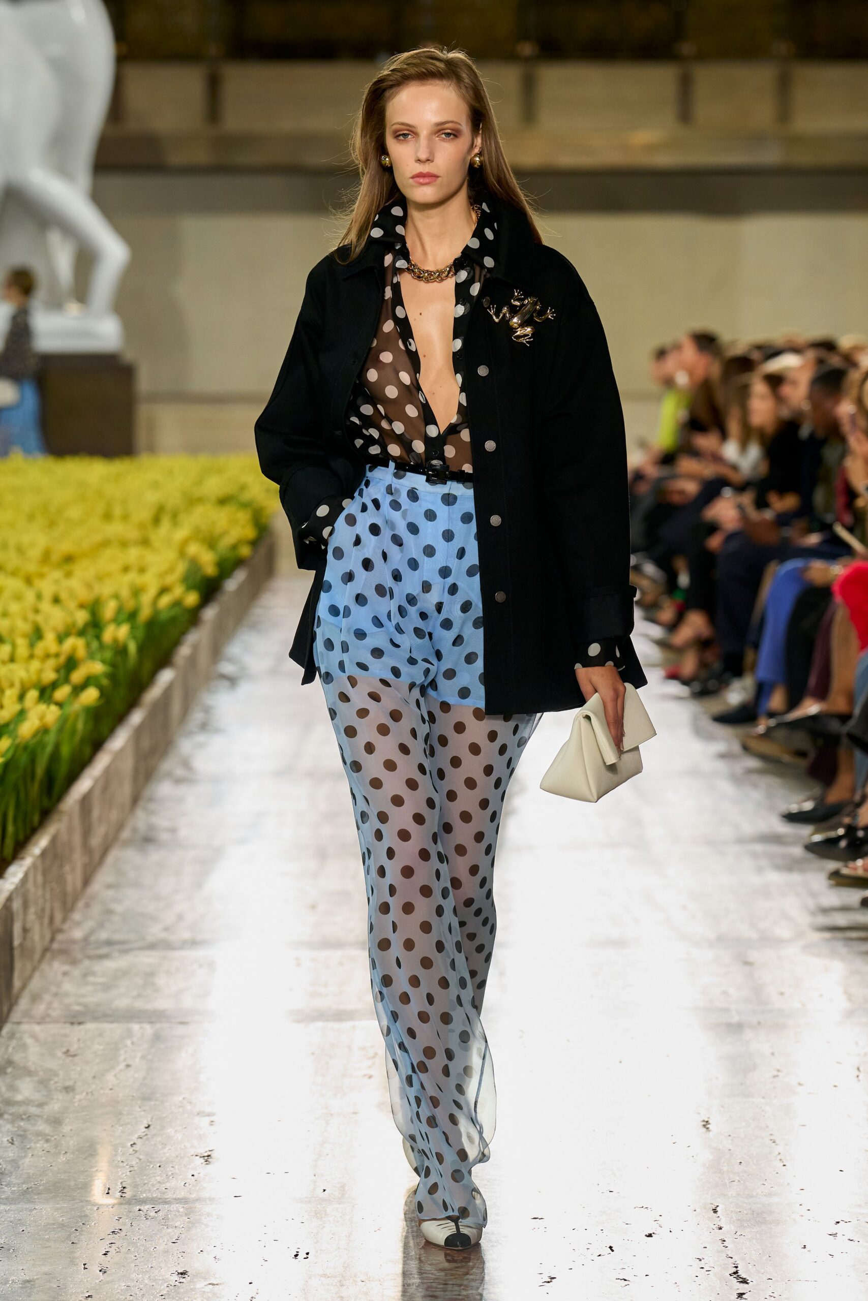 Black wool jacket with dull silver snaps and a frog brooch. Sheer black polka-dot chiffon blouse, plunged open to the waist. Powder-blue mesh trousers scattered with oversized black dots. Soft white folded clutch. Cream pumps with a black cap toe. Crossing a sculpture court after rain, flowers boxed neatly at the edge.