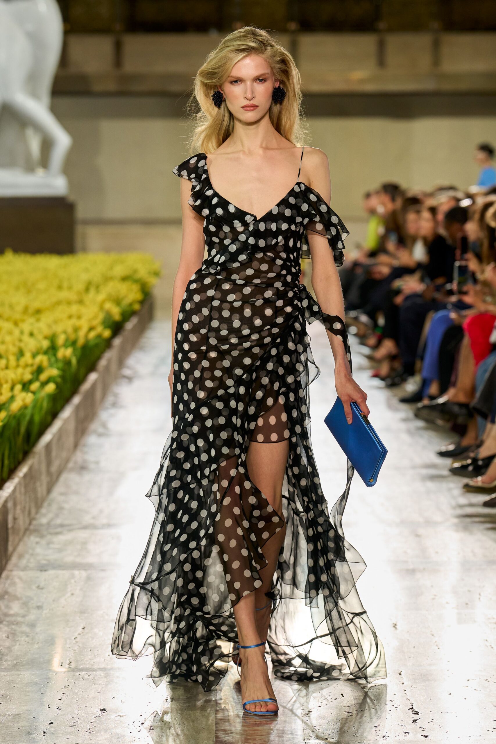 Sheer black chiffon gown in oversized white polka dots, ruffled off one shoulder. Flat cobalt clutch tucked under one hand. Bare blue ankle-strap sandals. Black clustered bead earrings. Walking beside a formal garden while the fountains are switched off.