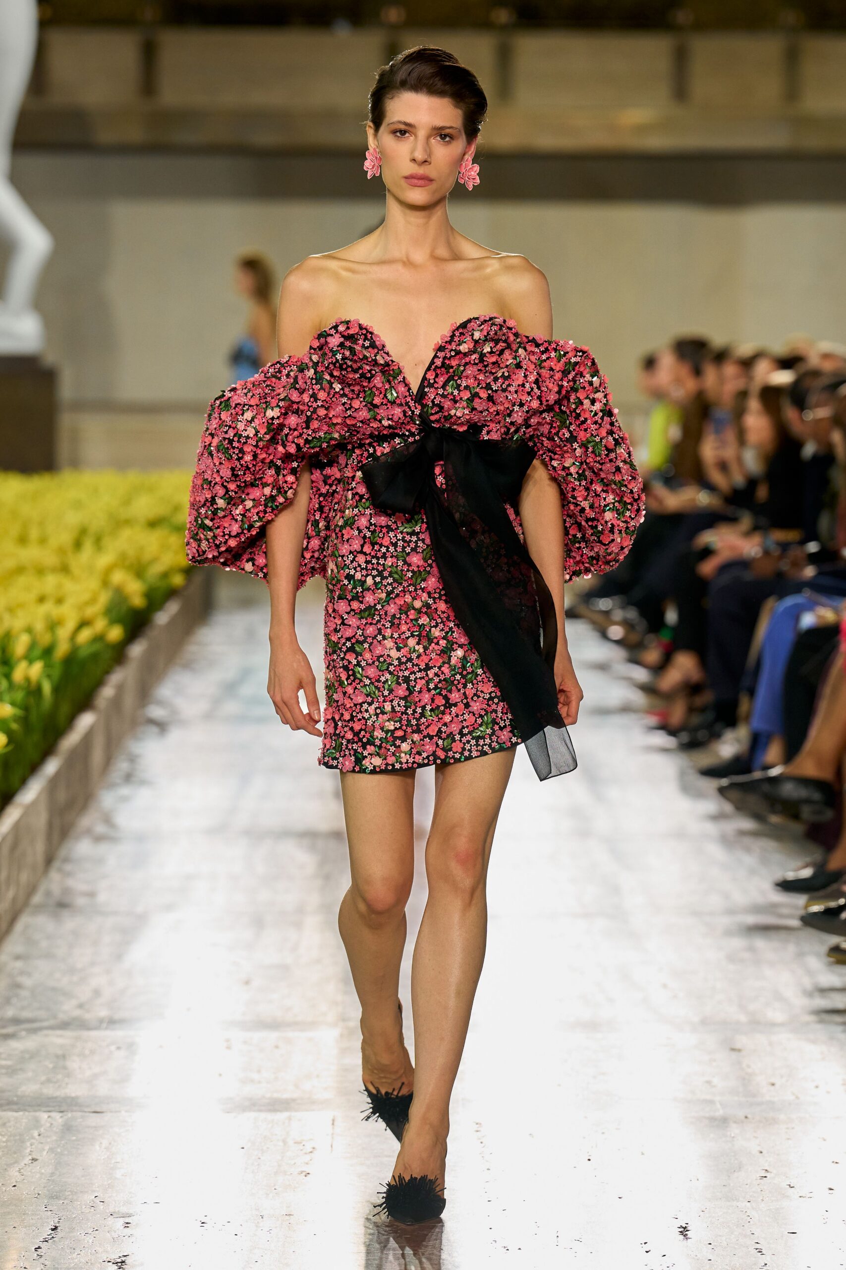 Pink flower-cluster drop earrings. Black mini dress packed with pink ditsy florals and blown-out off-shoulder poufs. Sheer black sash knotted at the bust, trailing past the hem. Black pumps tipped with spiky fringe pompons. Walking beside formal tulip beds just after the sprinklers shut off.